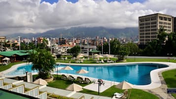 A swimming pool with lounge chairs and umbrellas, surrounded by greenery and a cityscape with mountains in the background.