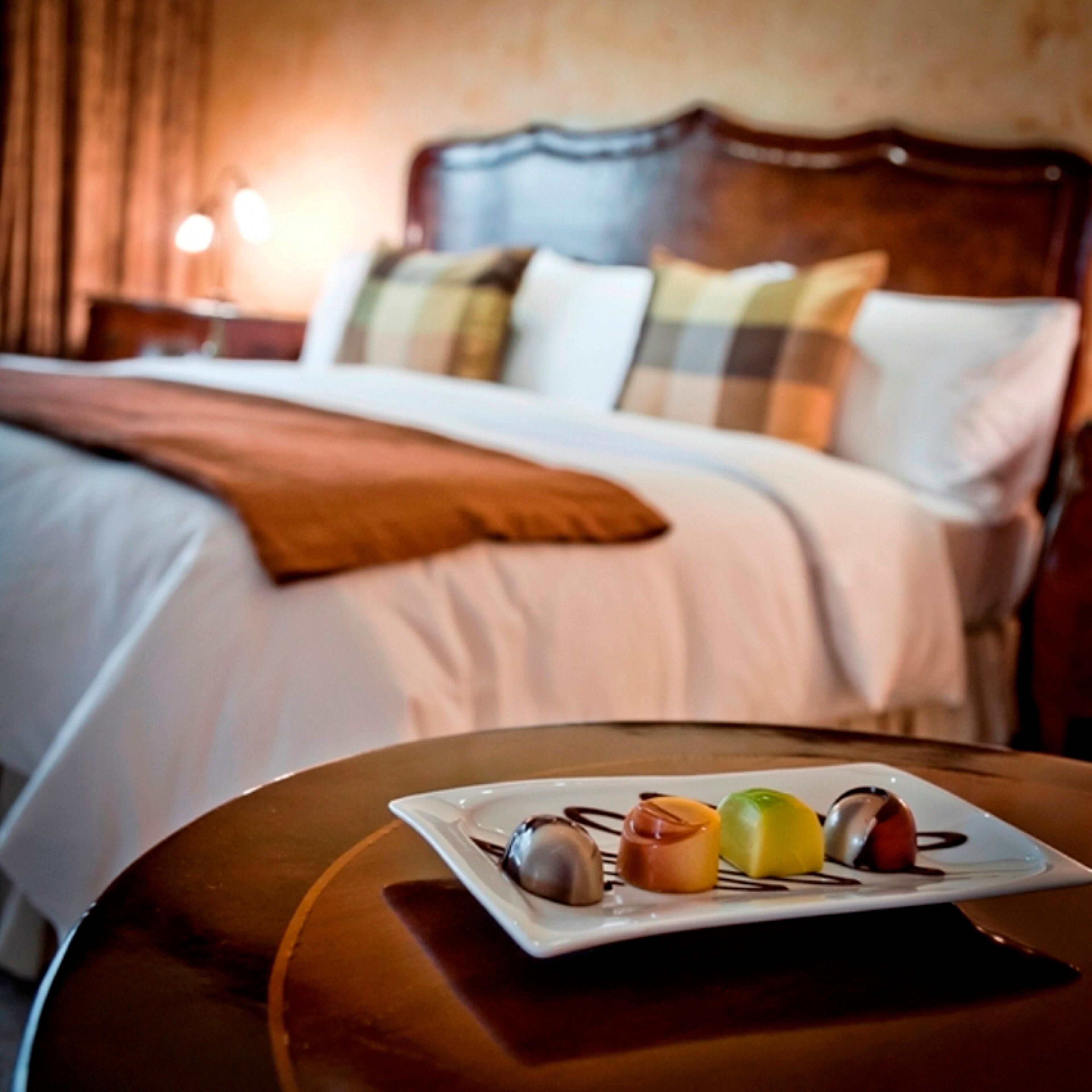 A neatly made bed with a brown throw blanket, a bedside table with a plate of dessert, and a nightstand with a lamp. - Hotel Tamanaco Caracas (Caracas)