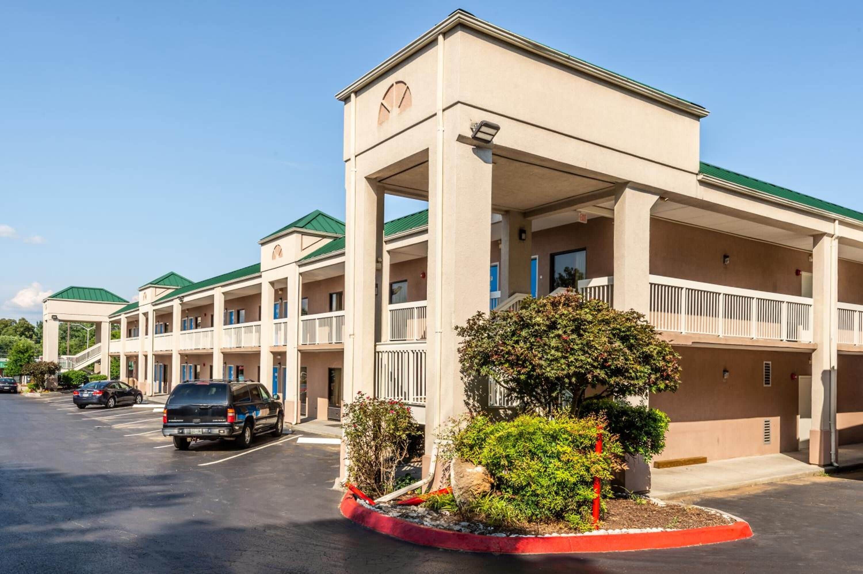 A multi-story motel with a central atrium, outdoor parking, and a landscaped area with shrubs and a small tree.
