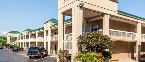 Un motel de varios pisos con un atrio central, estacionamiento exterior y un área ajardinada con arbustos y un árbol pequeño.