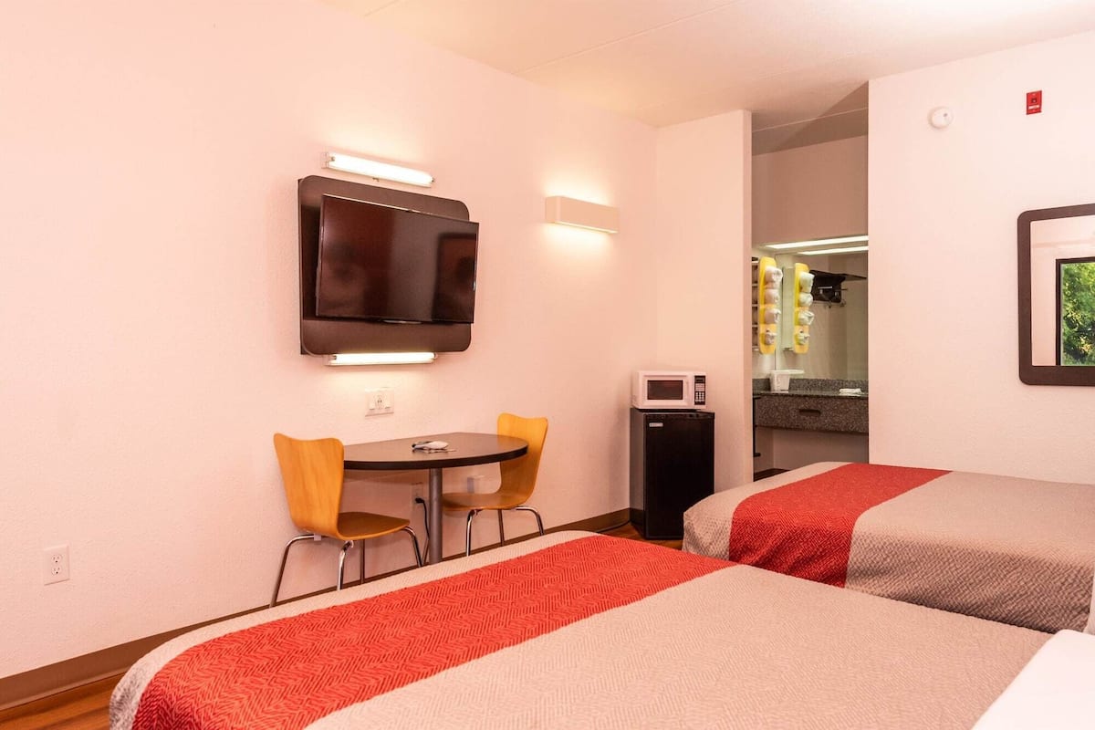 A hotel room with two beds, a small table with chairs, a TV mounted on the wall, a microwave, and a bathroom visible through an open door.