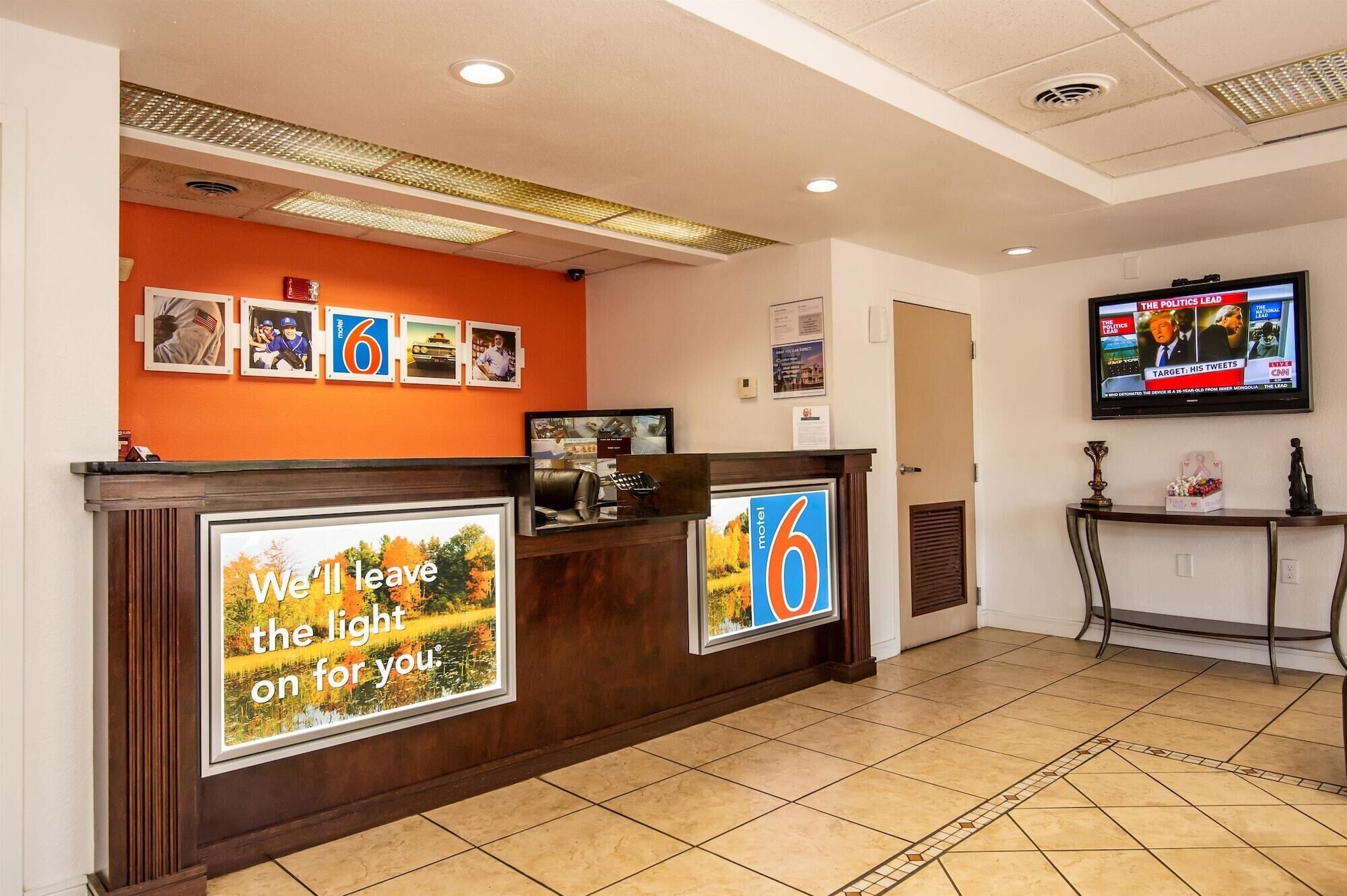 A hotel reception area with a wooden reception desk, a television screen displaying news, and a wall with framed pictures and a sign.