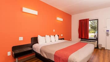 Hotellihuone, jossa on oranssi seinä, valkoisilla lakanoiden peitetty sänky, punavalkoinen peitto, pieni yöpöytä ja punaisella verholla varustettu ikkuna.