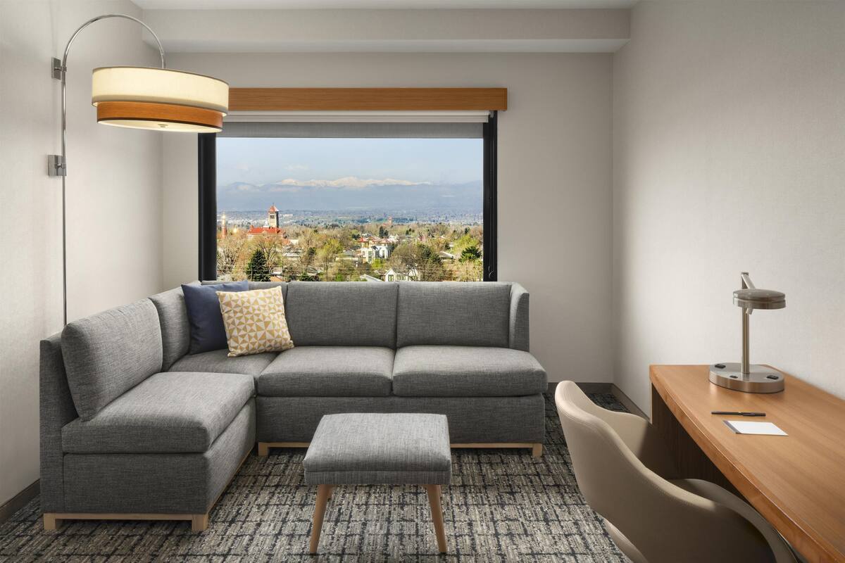 A modern hotel room with a gray sectional sofa, a small ottoman, a wooden desk, and a large window offering a city view.