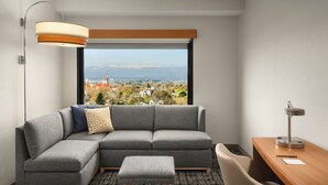 A modern room with a sectional sofa, a small ottoman, a desk, and a view of a cityscape through a large window. - Hyatt Place Denver/Cherry Creek (Glendale)