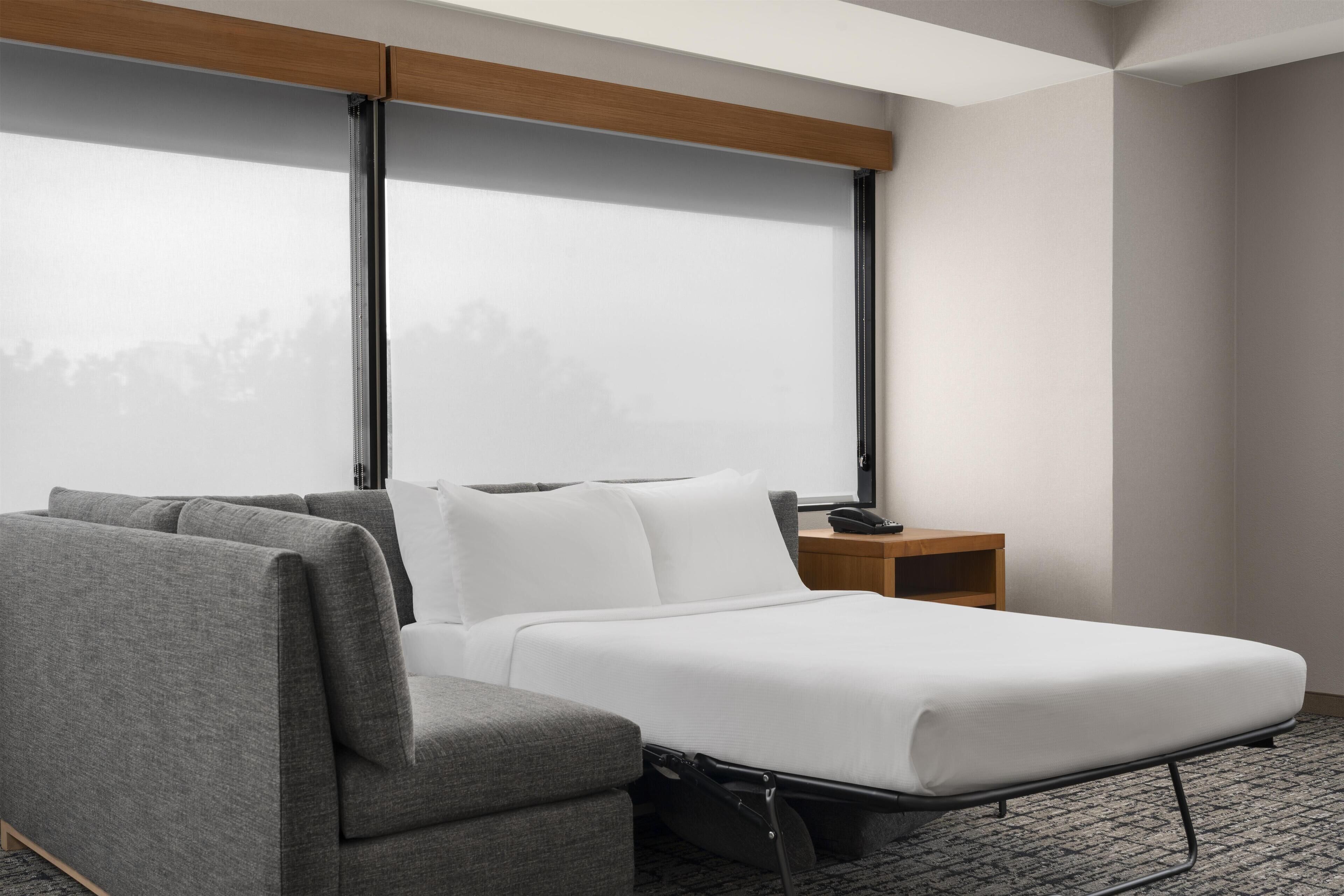 Quarto de hotel moderno com uma cama, um sofá cinzento, uma mesinha de madeira e uma grande janela com vista para árvores.