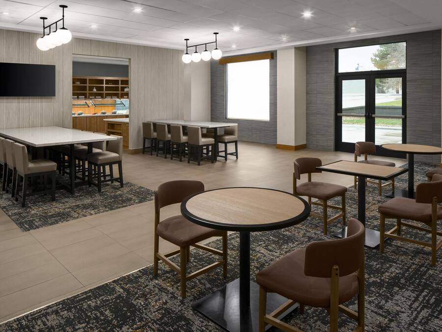 A spacious hotel dining area with multiple tables and chairs, a flat-screen TV mounted on the wall, and large windows providing natural light.
