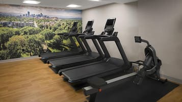 A gym with treadmills and an elliptical machine, a cityscape mural on the wall, and wooden flooring.