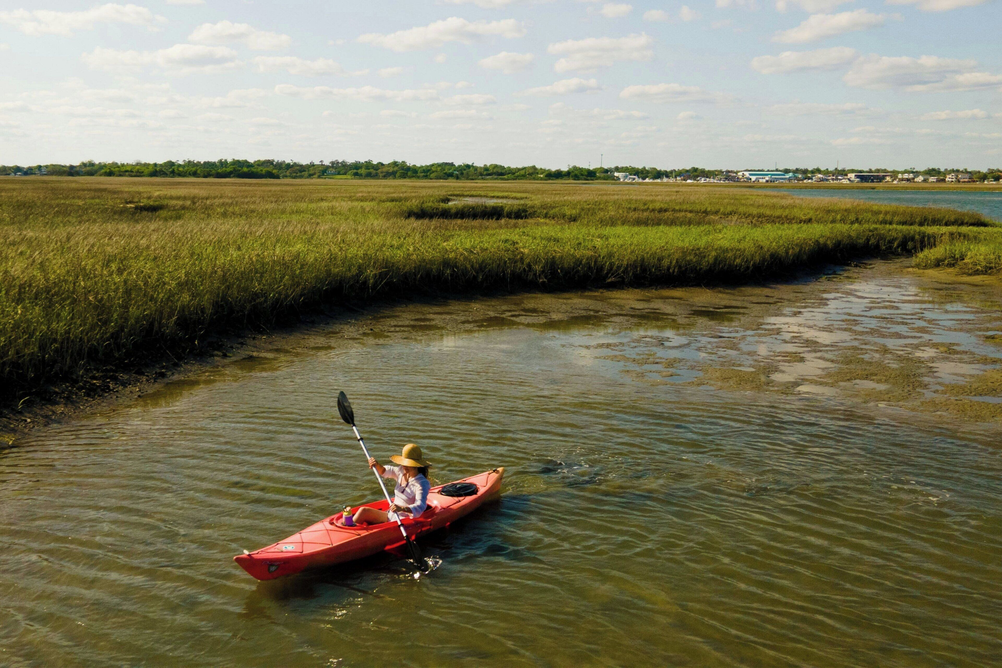 Une personne dans un kayak rouge, sur une rivière paisible, entourée de zones marécageuses.