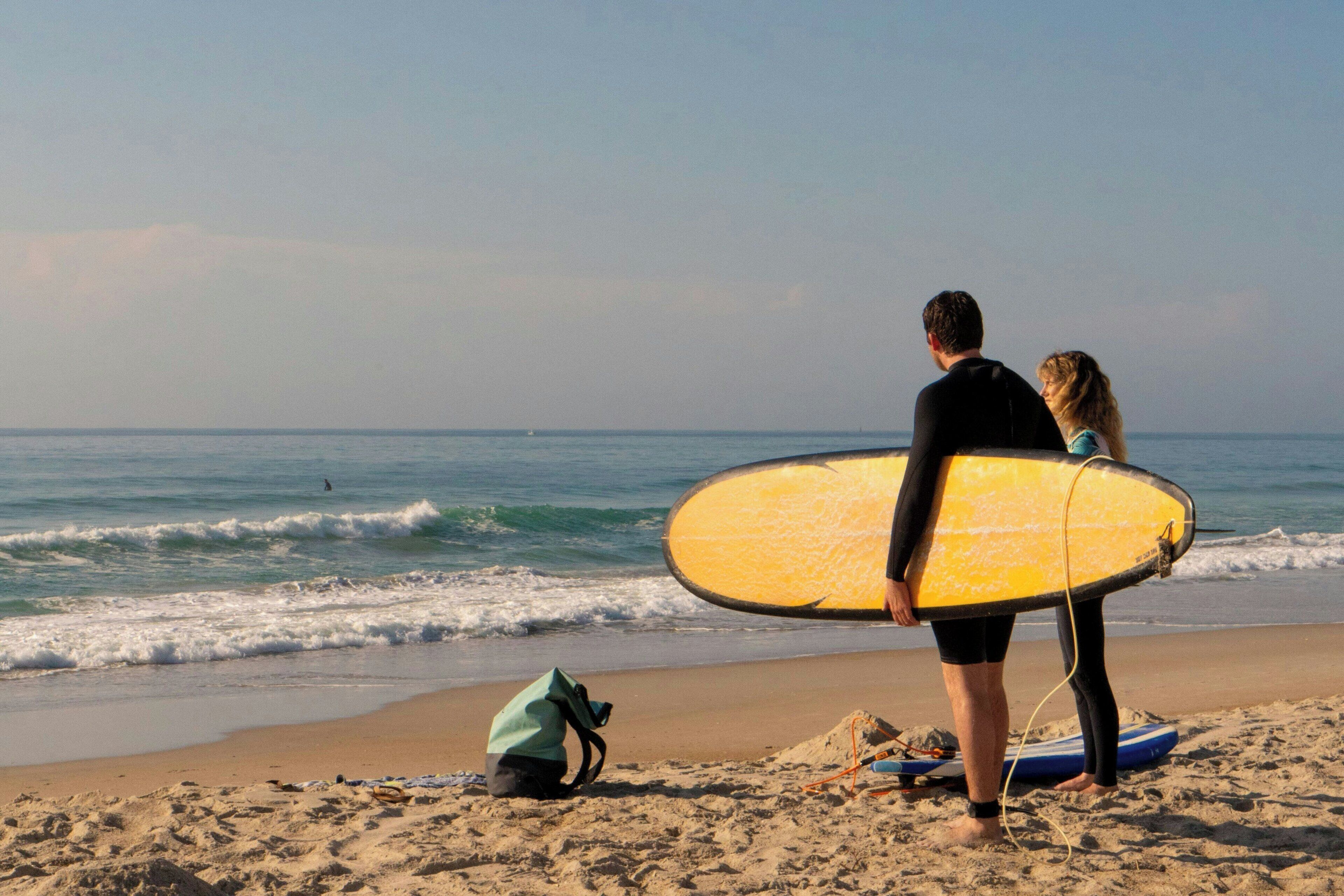 A beach scene with a person holding a yellow surfboard, another person standing beside them, and a green bag on the sand.