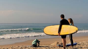 Una spiaggia con una persona che tiene un surf giallo, un'altra persona accanto e una borsa verde sulla sabbia.