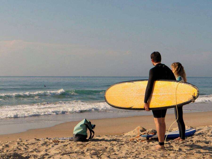Una spiaggia con una persona che tiene un surf giallo, un'altra persona accanto e una borsa verde sulla sabbia.