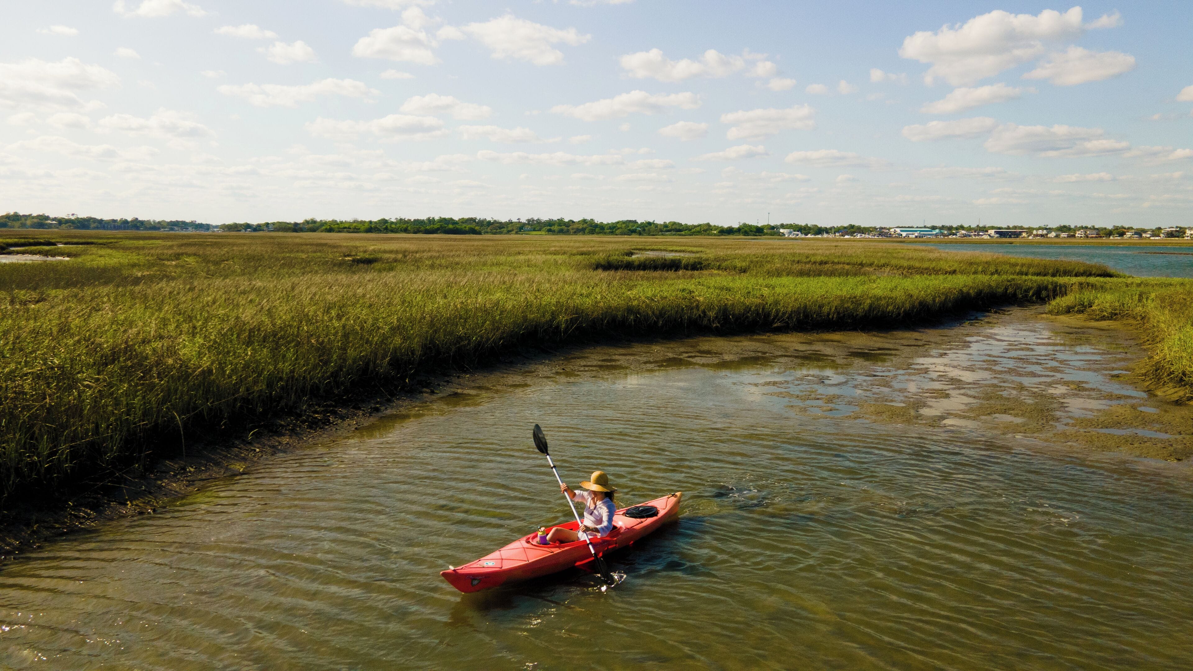 Un kayakiste dans un kayak rouge, sur une rivière paisible, entouré de marais.