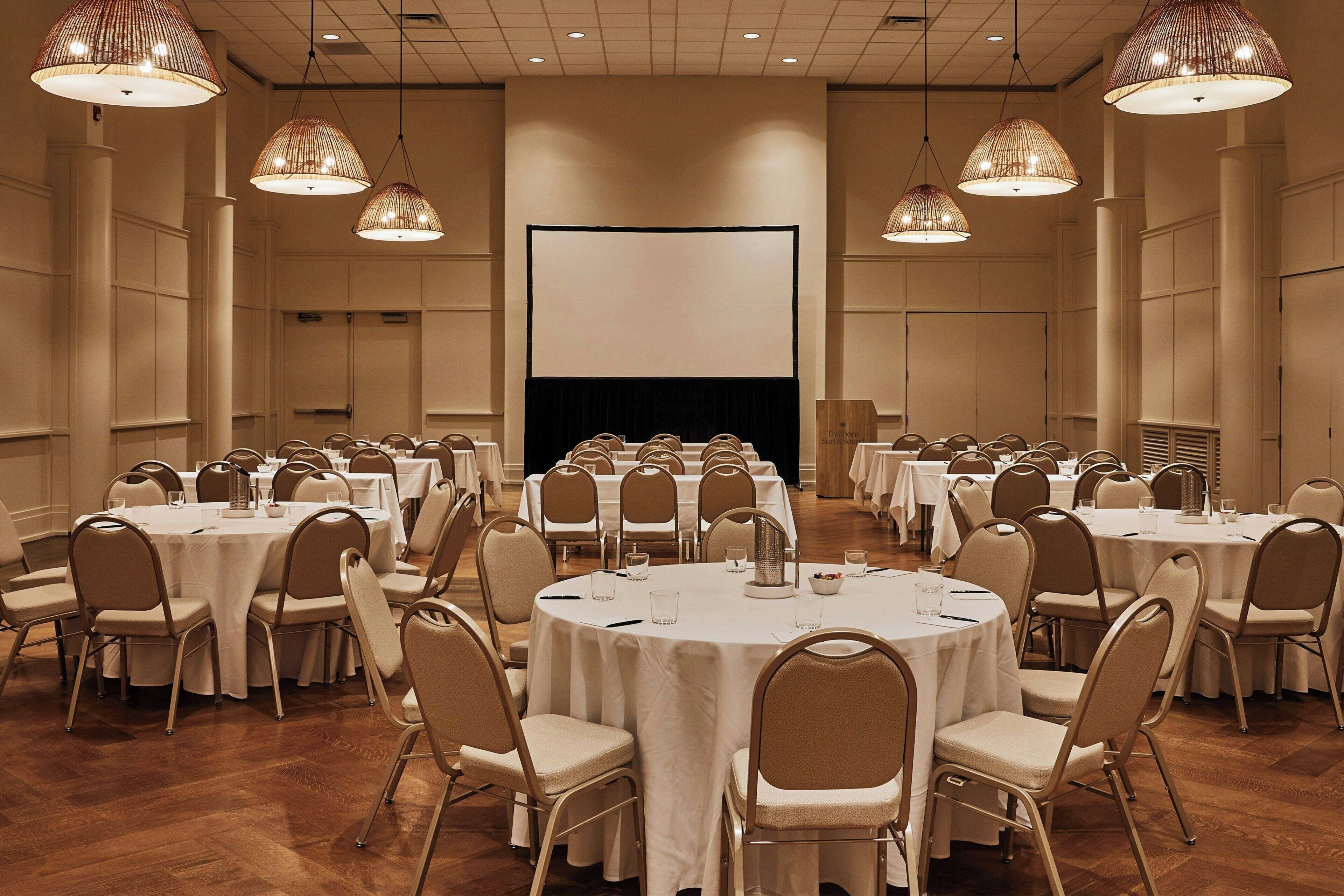 A conference room with multiple round tables, chairs, and large hanging lamps.