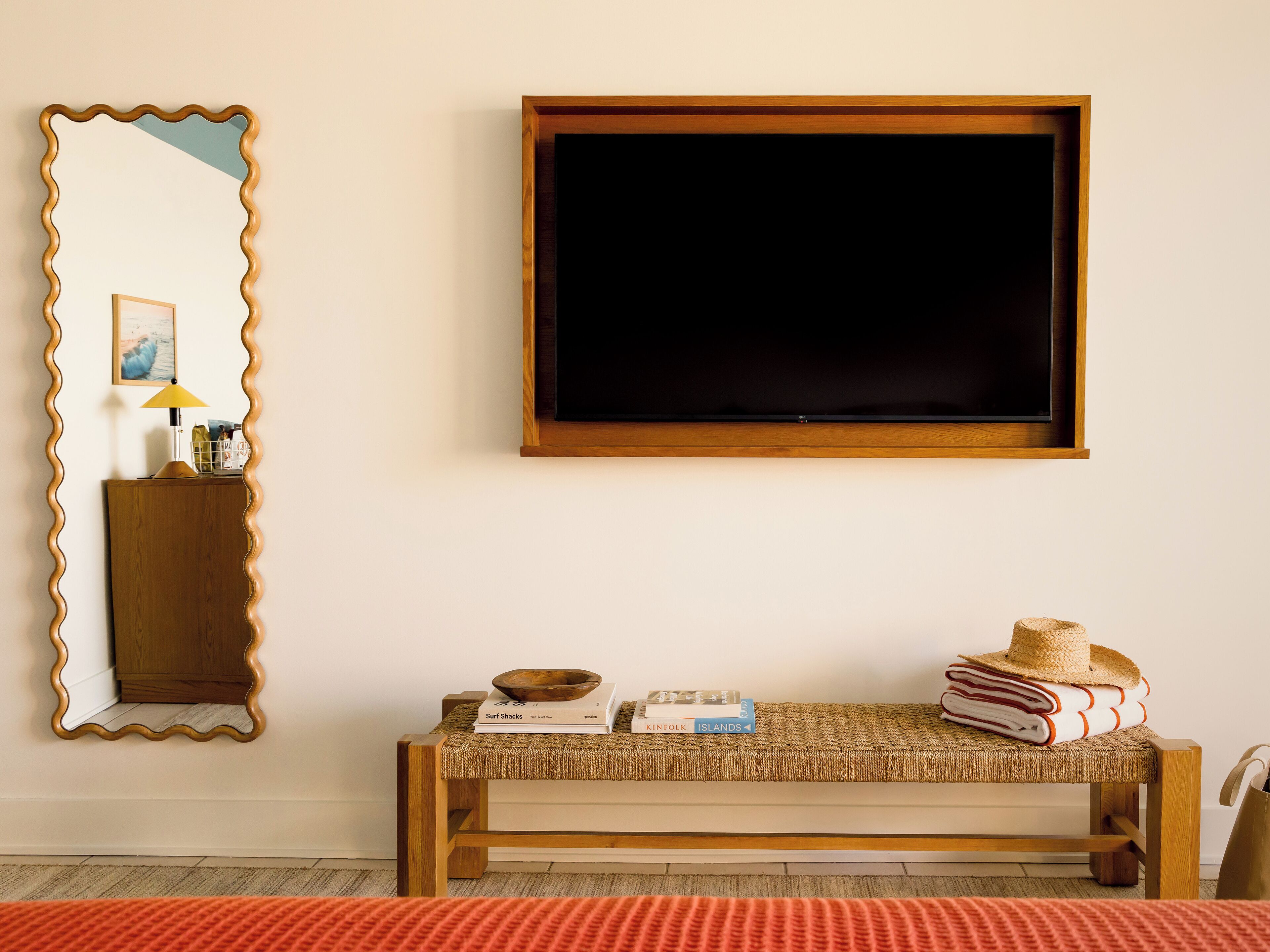 A room with a wooden bench, a mirror, a television, and a small table with books and a hat.