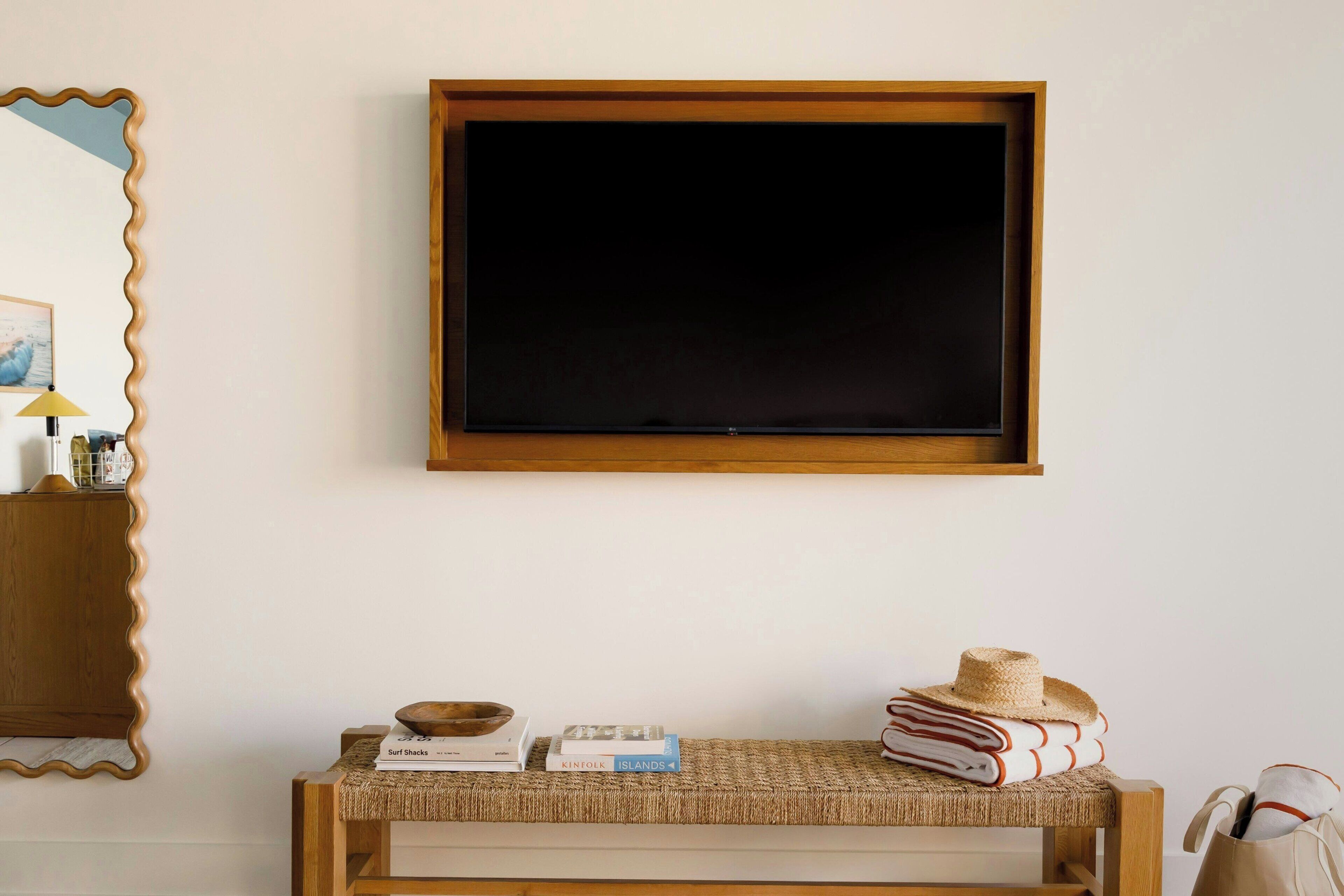 A room with a wooden-framed television mounted on the wall, a woven bench with books and a hat, and a wooden side table with a lamp.