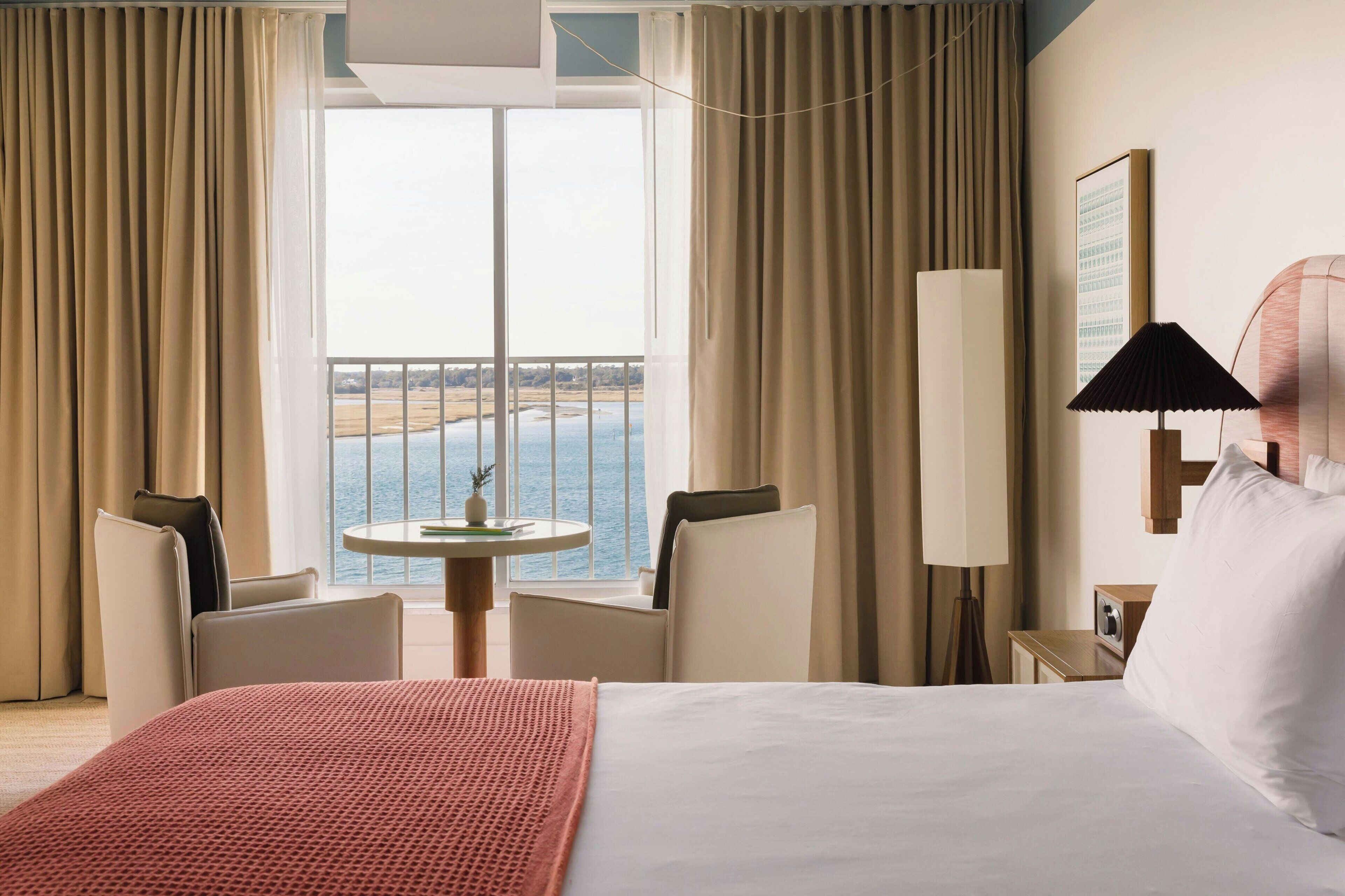 A hotel room with a bed, a table, two chairs, a lamp, and a view of the sea.