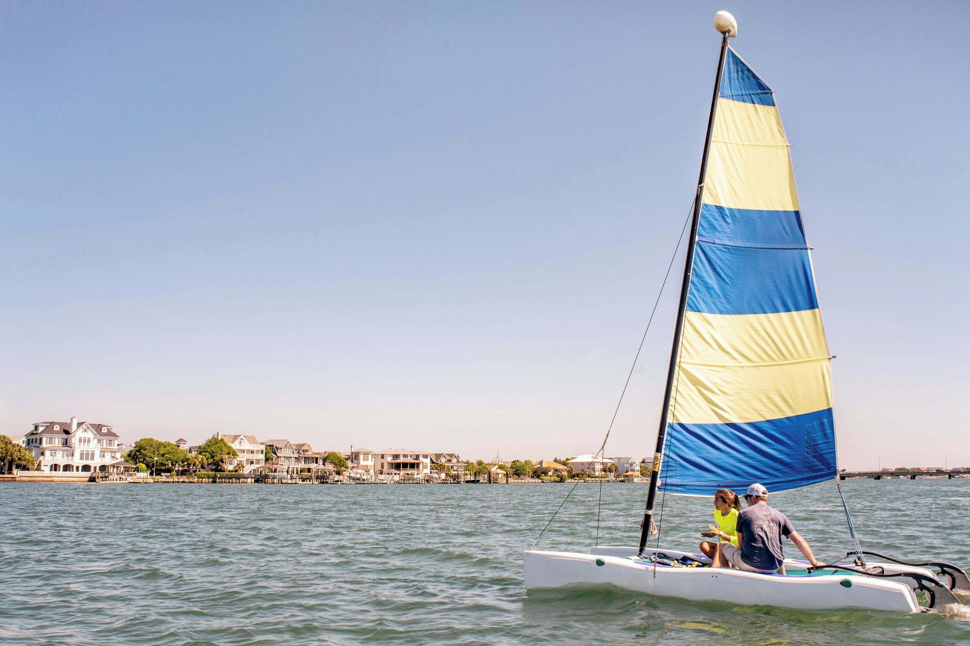 A sailboat with a blue and yellow sail, two people on board, and a waterfront with houses in the background.