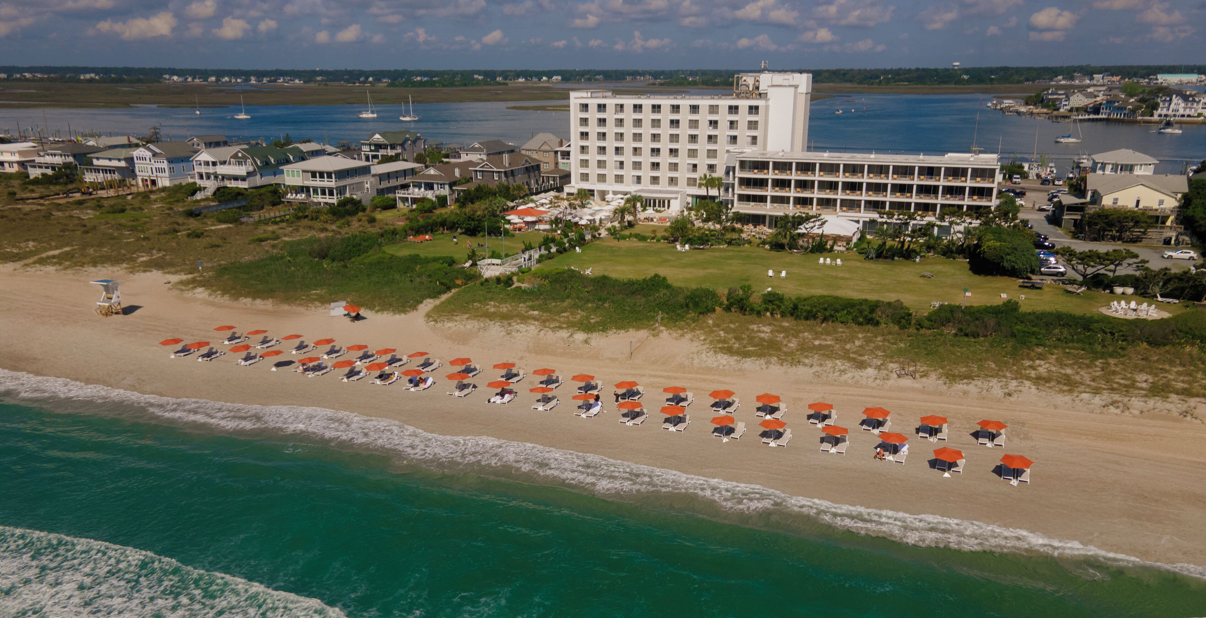 An aerial view of a coastal hotel with a beachfront, rows of beach chairs, and a large building with multiple floors.