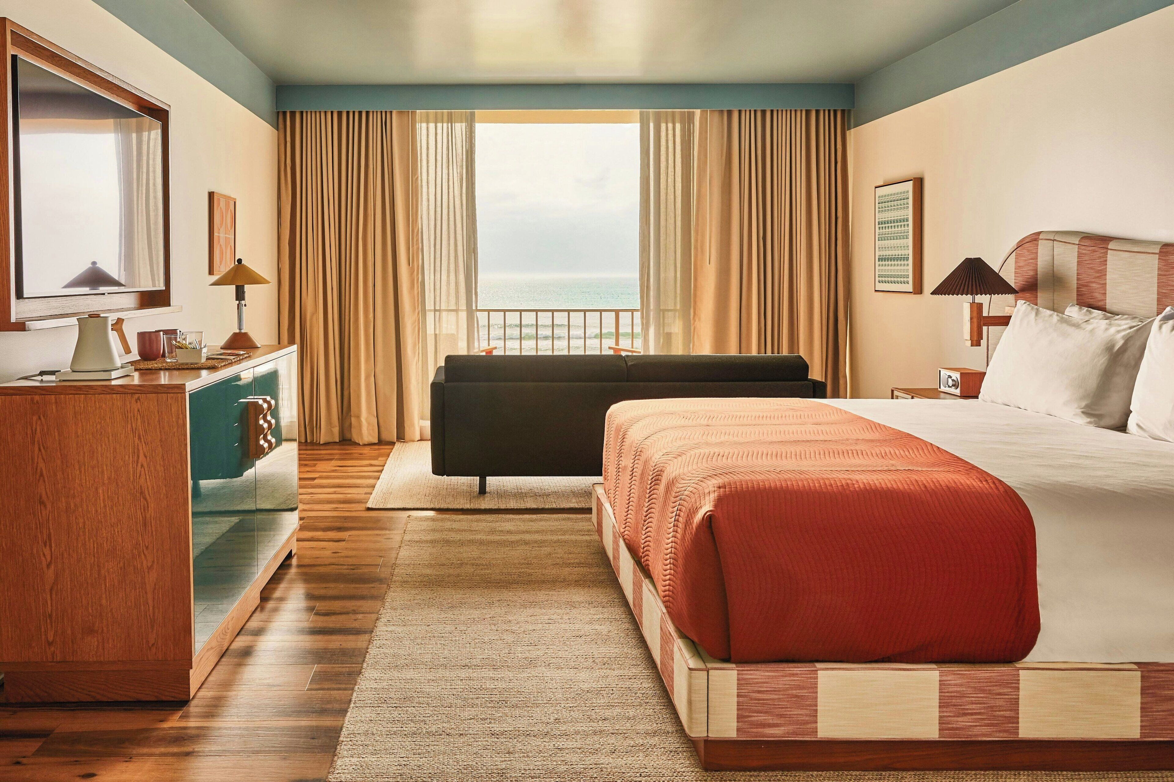 Habitación de hotel con una cama grande, un sofá, un escritorio con lámpara, vista al mar y una ventana con cortinas.