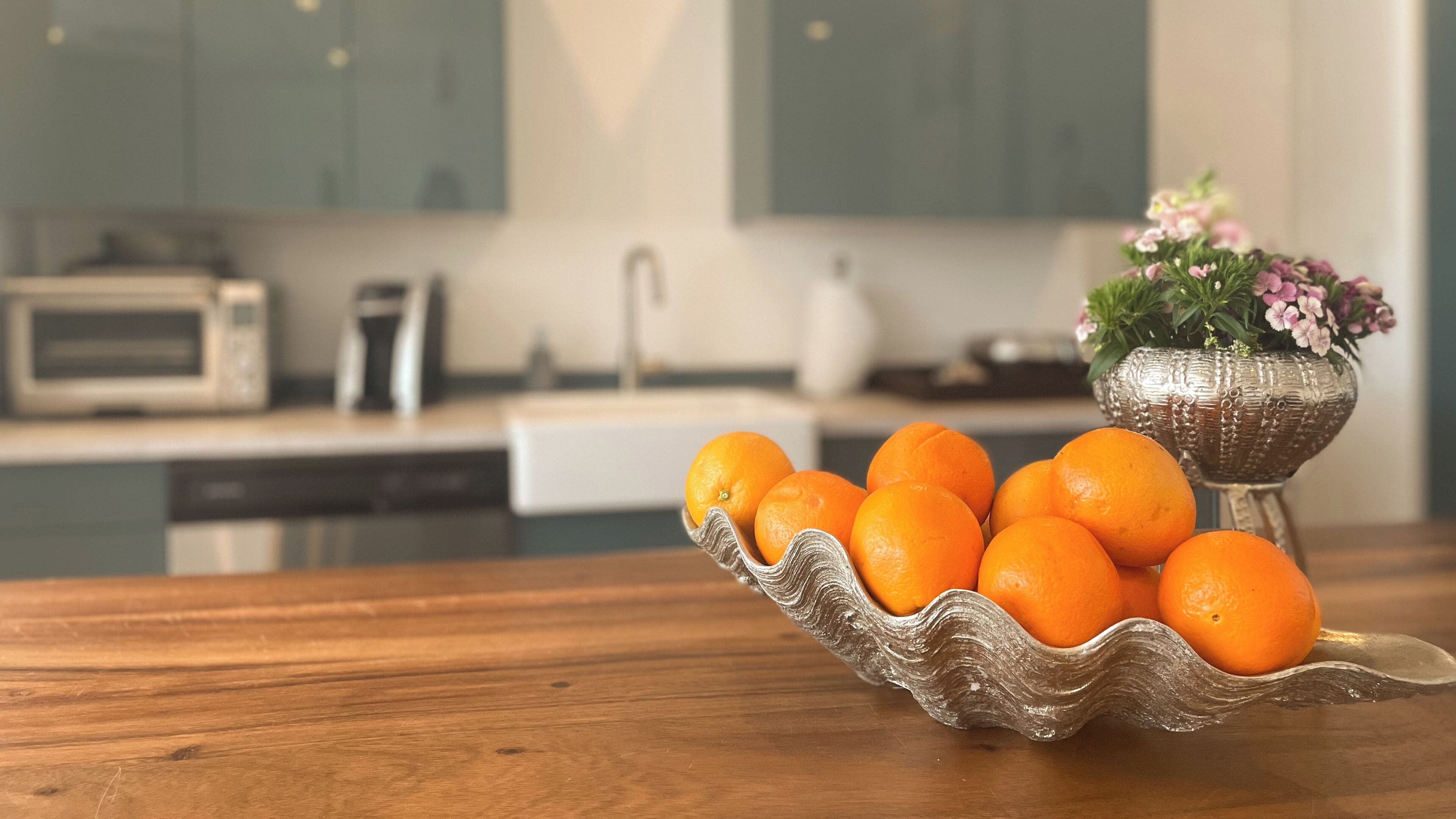 A kitchen with a wooden table, a bowl of oranges, a microwave, and a decorative vase with flowers.
