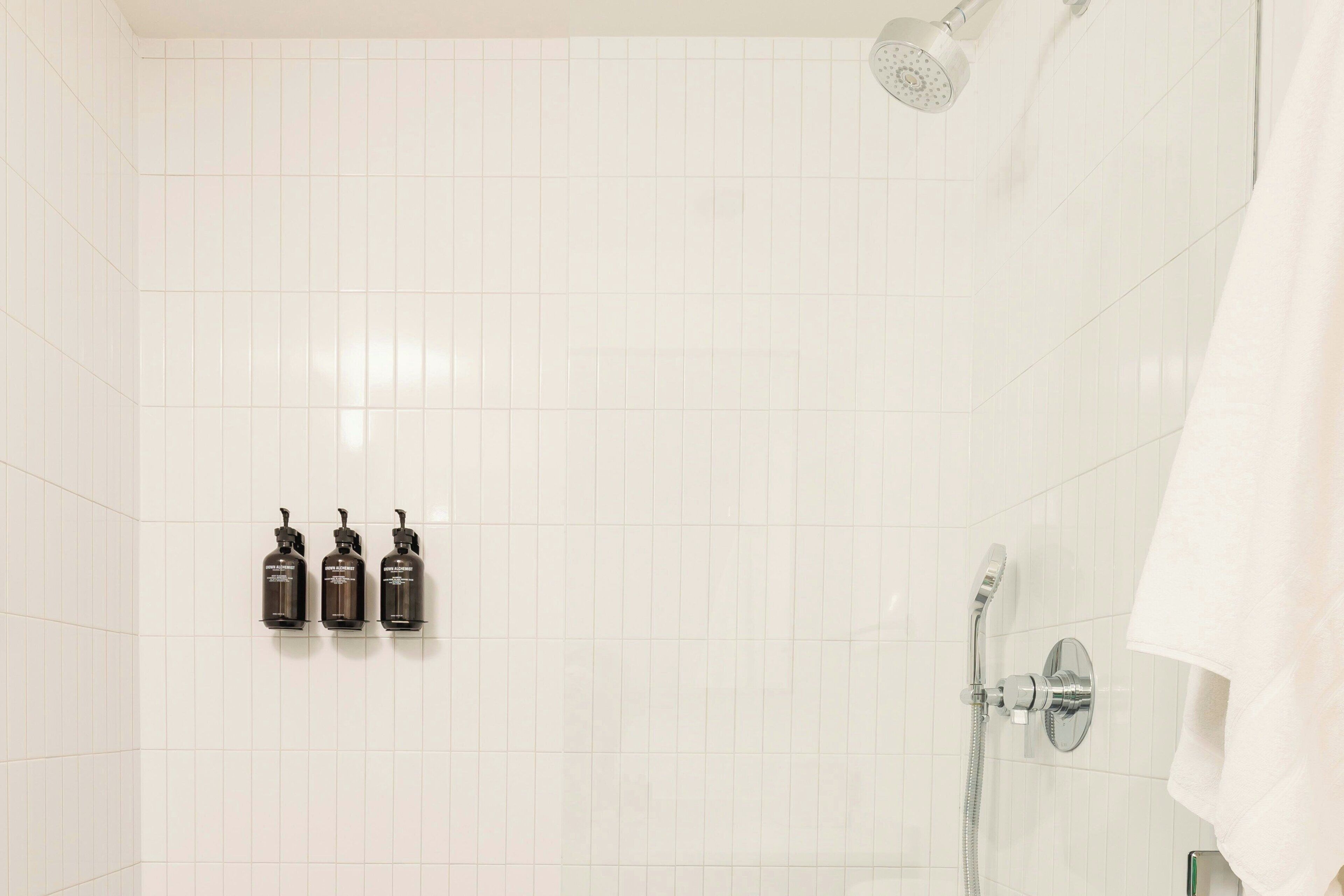 Ein Badezimmer mit weißen Fliesen, einer Dusche inklusive Handbrause und drei Flaschen Shampoo oder Conditioner an der Wand.