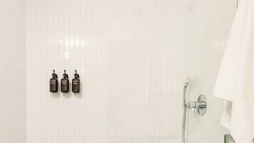 Un baño con paredes cubiertas de azulejos blancos, una ducha con cabezal de mano y tres botellas de champú o acondicionador en la pared.