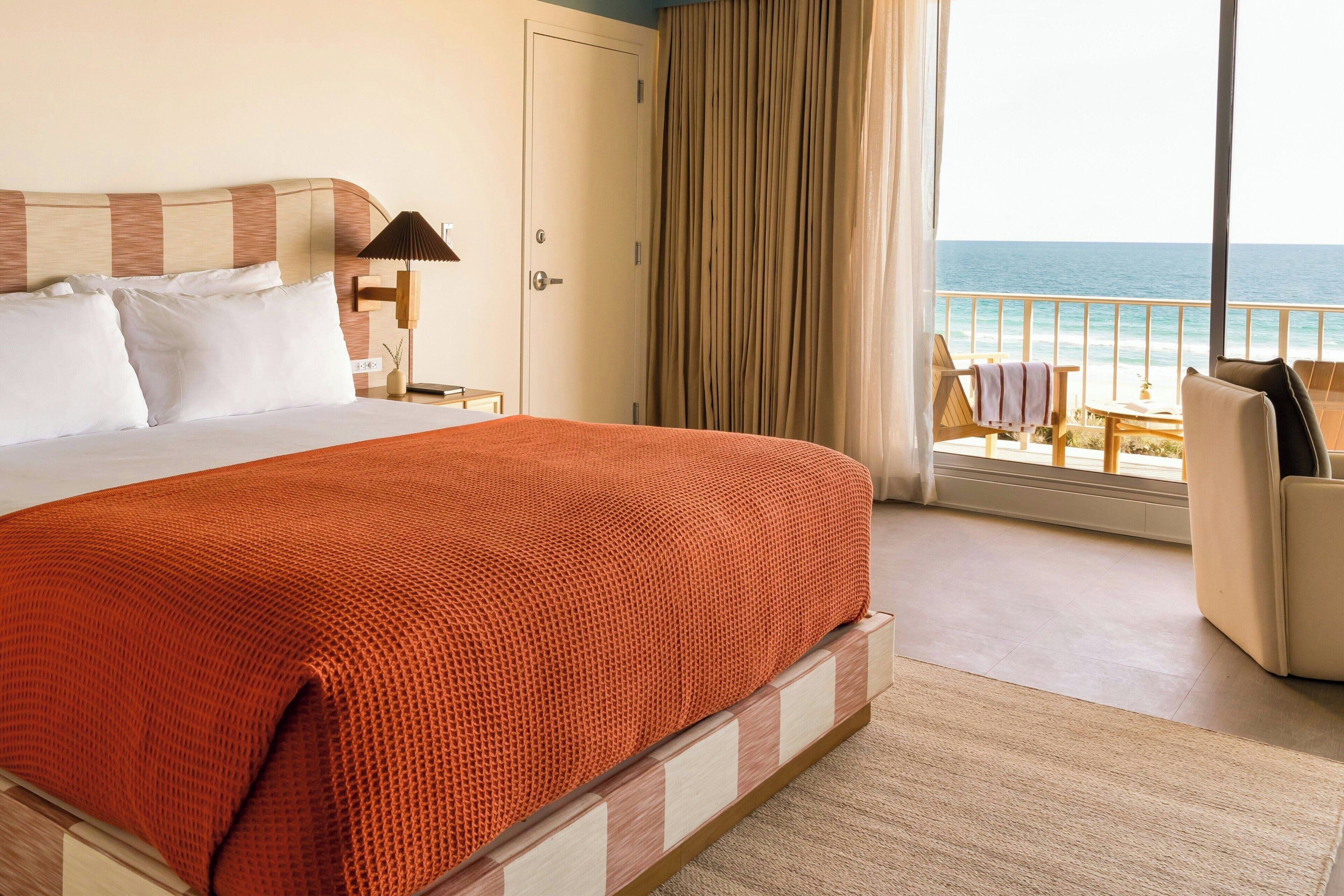 A hotel room with a large bed, a nightstand, a lamp, a view of the ocean, and a balcony with seating.