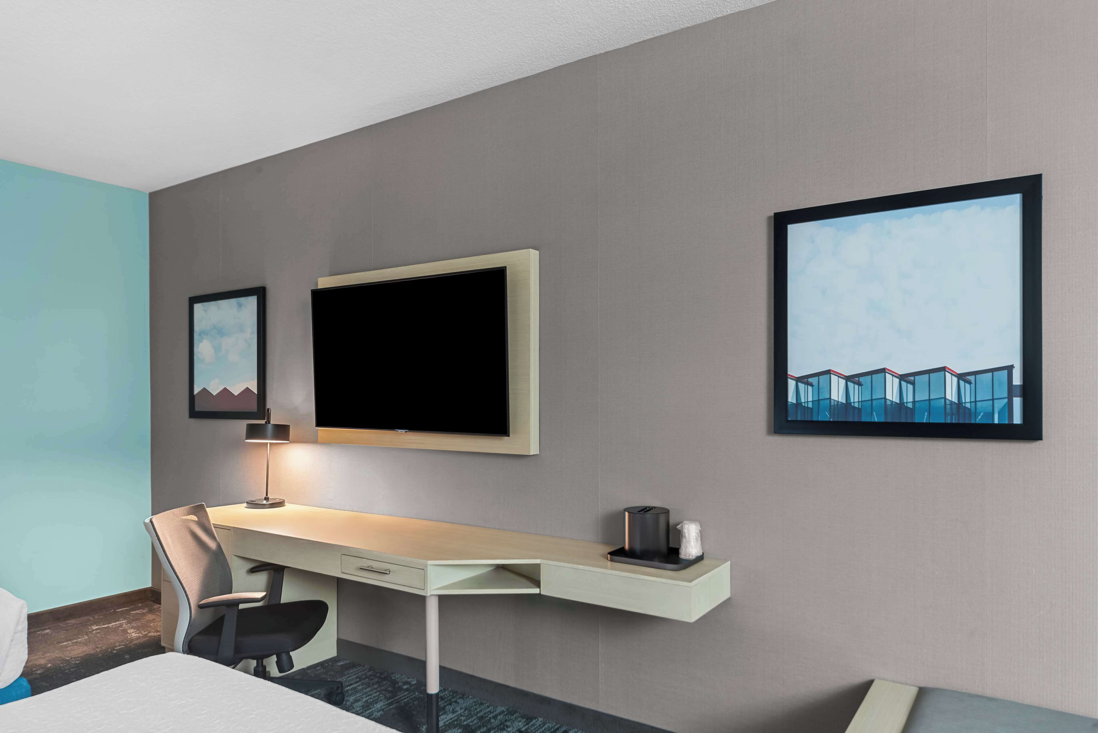 A hotel room with a bed, a desk with a chair, a flat-screen TV, and two framed pictures on the wall.