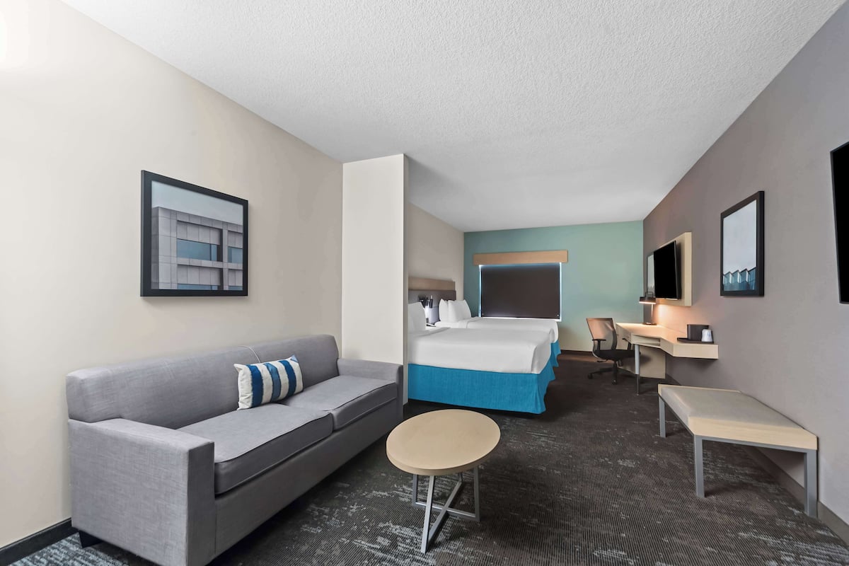 A hotel room with a gray sofa, a small round table, a bed with blue bedding, a desk with a chair, and a large mirror on the wall.