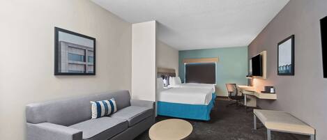 Quarto de hotel com sofá cinzento, mesa redonda pequena, cama com roupa de cama azul, escrivaninha com cadeira e um espelho grande na parede.