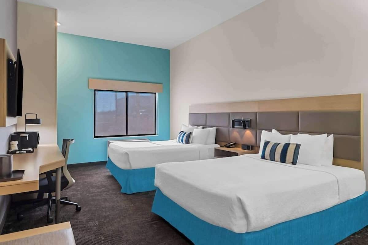 A hotel room with two beds, a desk, and a chair. There is a window with blinds, a flat-screen TV mounted on the wall, and a bedside table with a lamp.