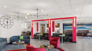 Eine moderne Lobby mit einer großen Uhr, roten Akzenten und verschiedenen Sitzbereichen, darunter Sofas und Sessel.