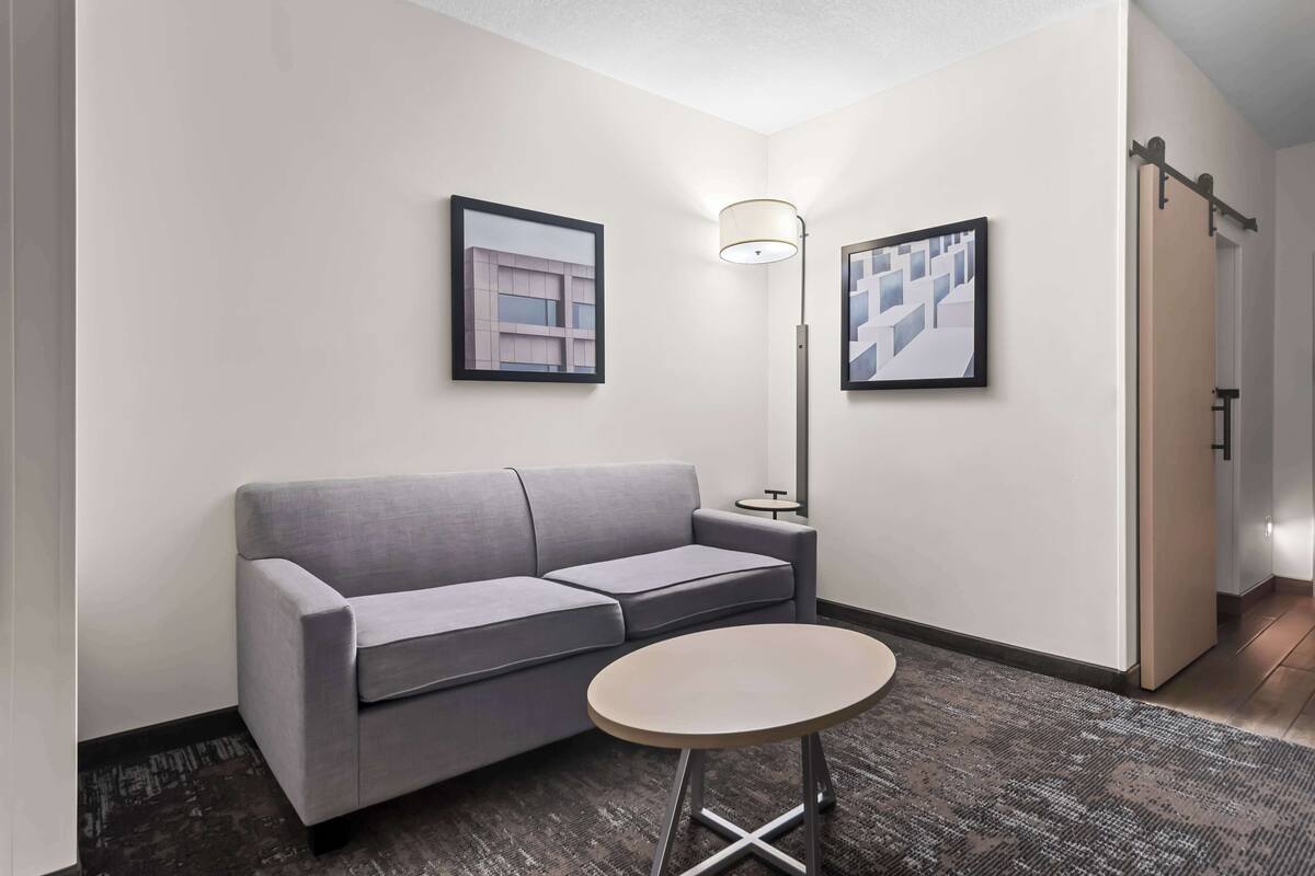 A modern hotel room with a grey sofa, a small round table, and two framed artworks on the walls.
