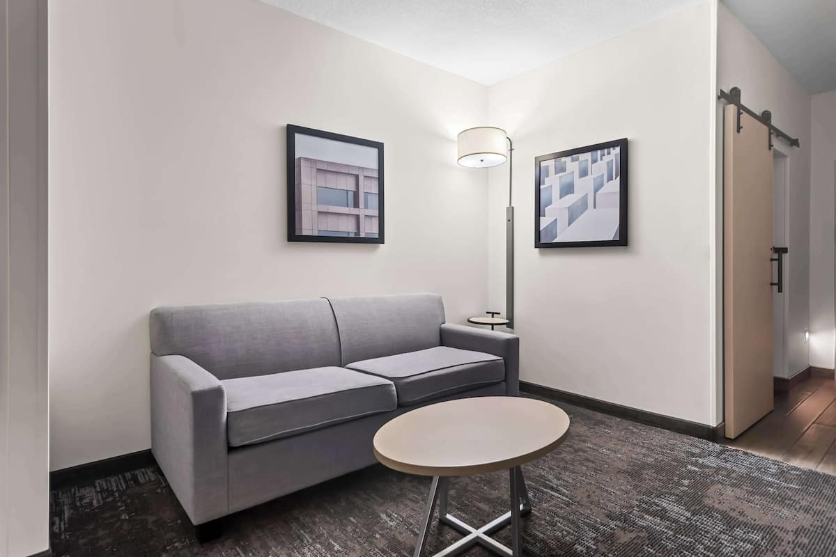 A modern hotel room with a grey sofa, a small round table, and two framed artworks on the walls.