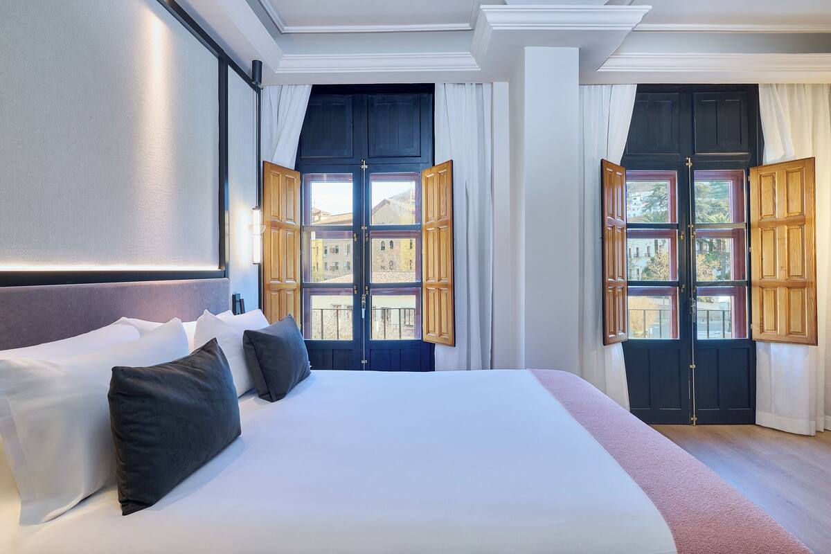Habitación de hotel con una cama grande, dos ventanas con contraventanas de madera, un cabecero y vistas a edificios.