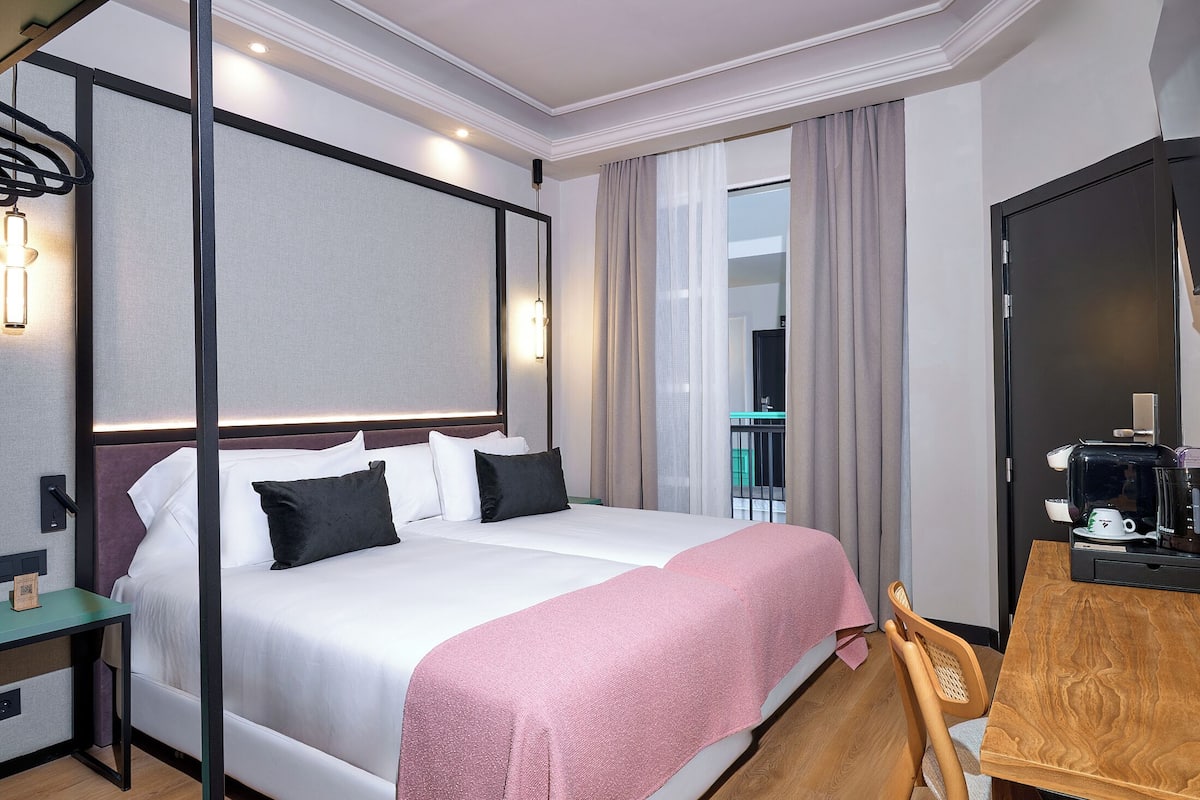 Habitación de hotel con cama, escritorio, silla, cafetera y vistas al exterior.