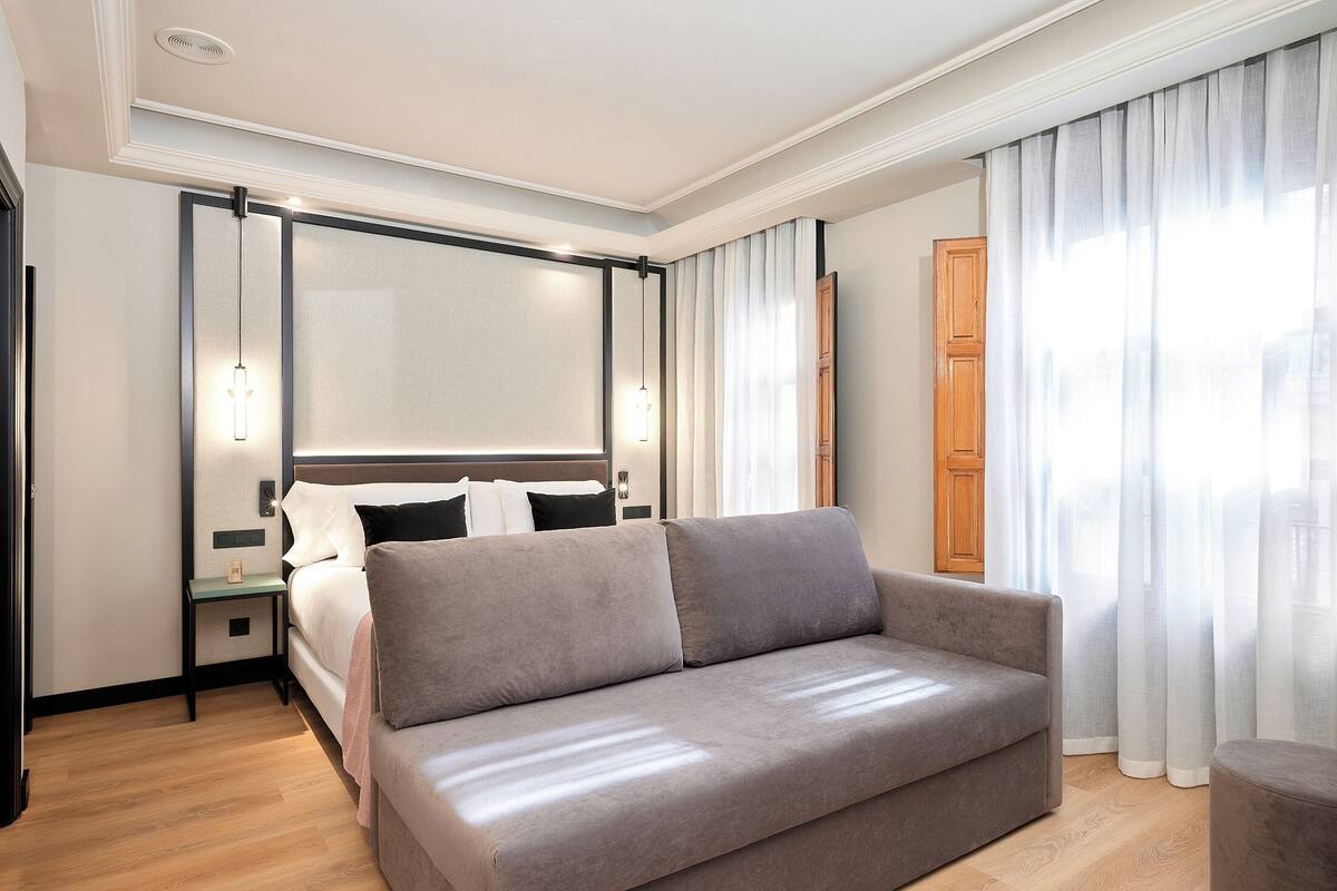 Una habitación de hotel moderna con cama, sofá, mesita de noche y un amplio ventanal con cortinas.