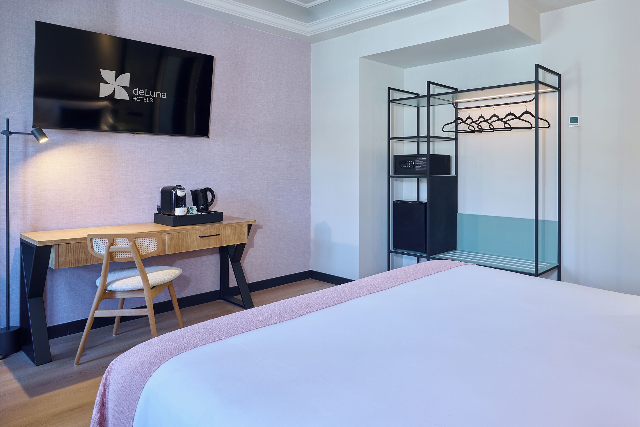 Habitación de hotel con cama, escritorio con silla, televisión, cafetera y perchero.