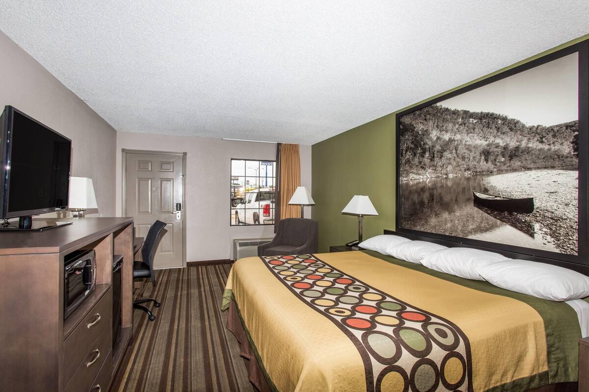Une chambre d’hôtel comprenant un lit, un bureau avec une télévision, une chaise, deux lampes et une grande fenêtre donnant sur un lac.