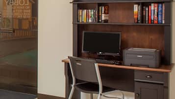 Un escritorio con monitor, teclado e impresora. Hay una estantería con varios libros y una silla frente al escritorio.