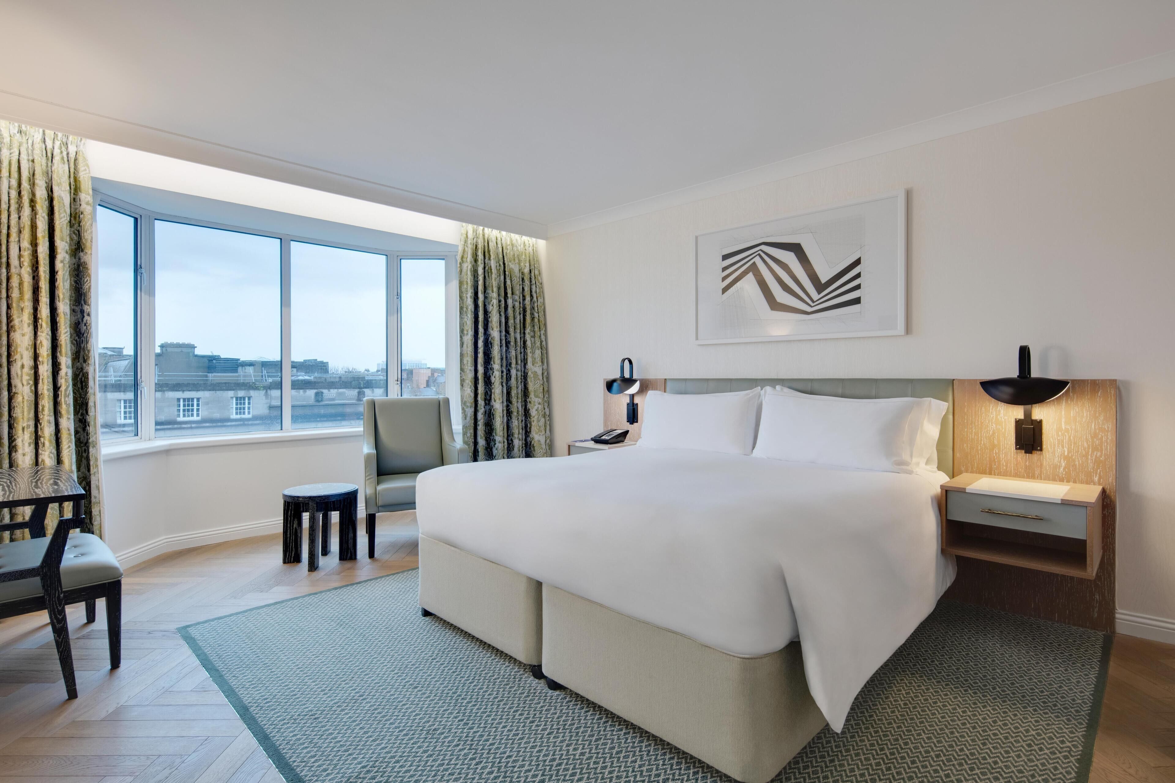 A hotel room with a large bed, a desk, a chair, a nightstand, and a view of the city through large windows.