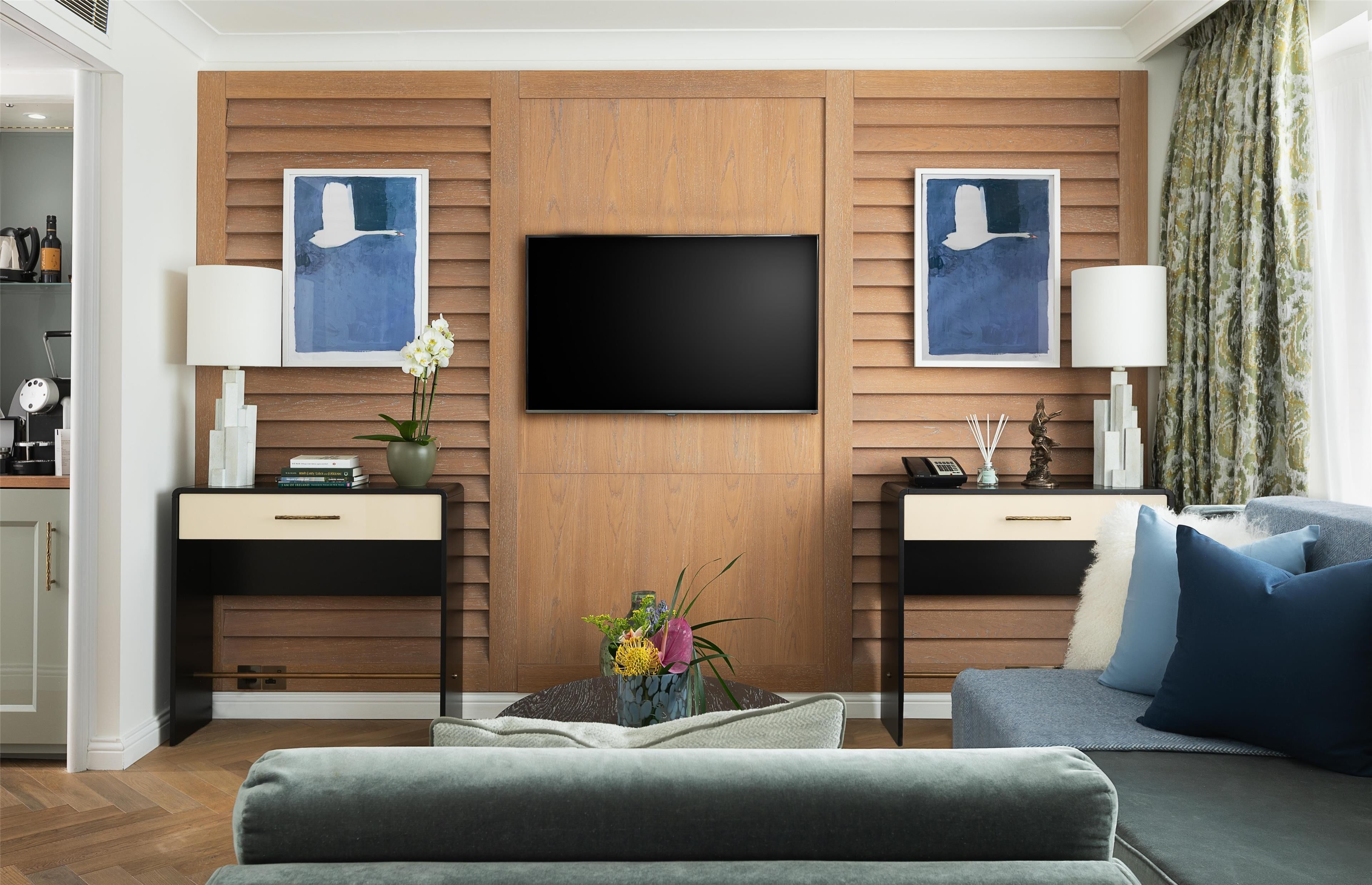 Una sala de estar moderna con un televisor de pantalla plana montado en una pared de madera, dos mesitas de noche con lámparas, un sofá y plantas decorativas.
