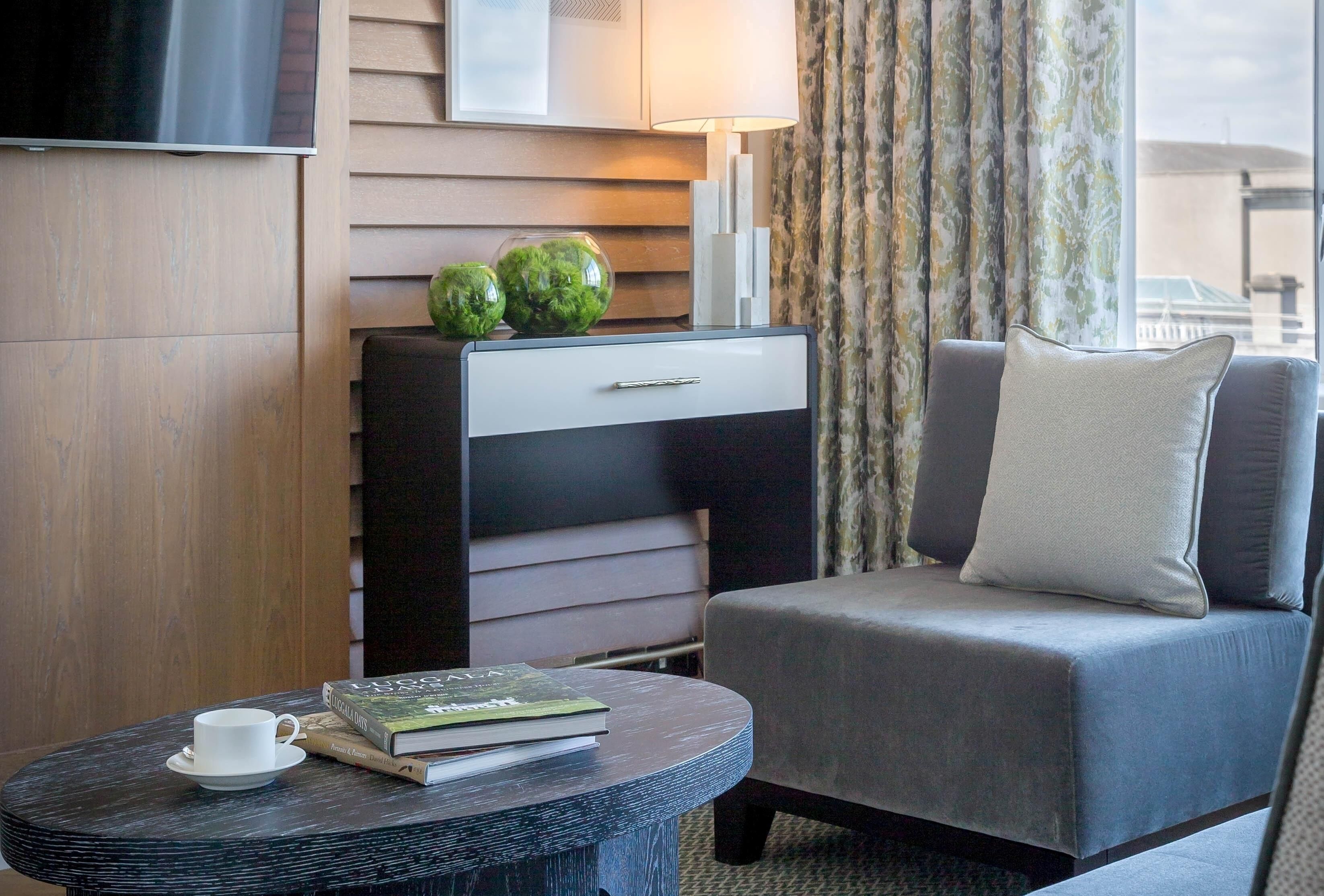 Une chambre d’hôtel avec un canapé, une petite table ronde sur laquelle se trouvent une tasse et un livre, une télévision et une plante décorative.