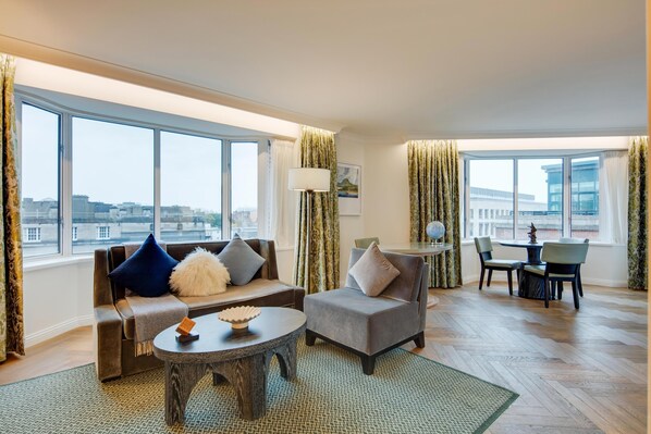 A spacious living room with a sofa, armchair, and round coffee table. Large windows offer a view of the cityscape.