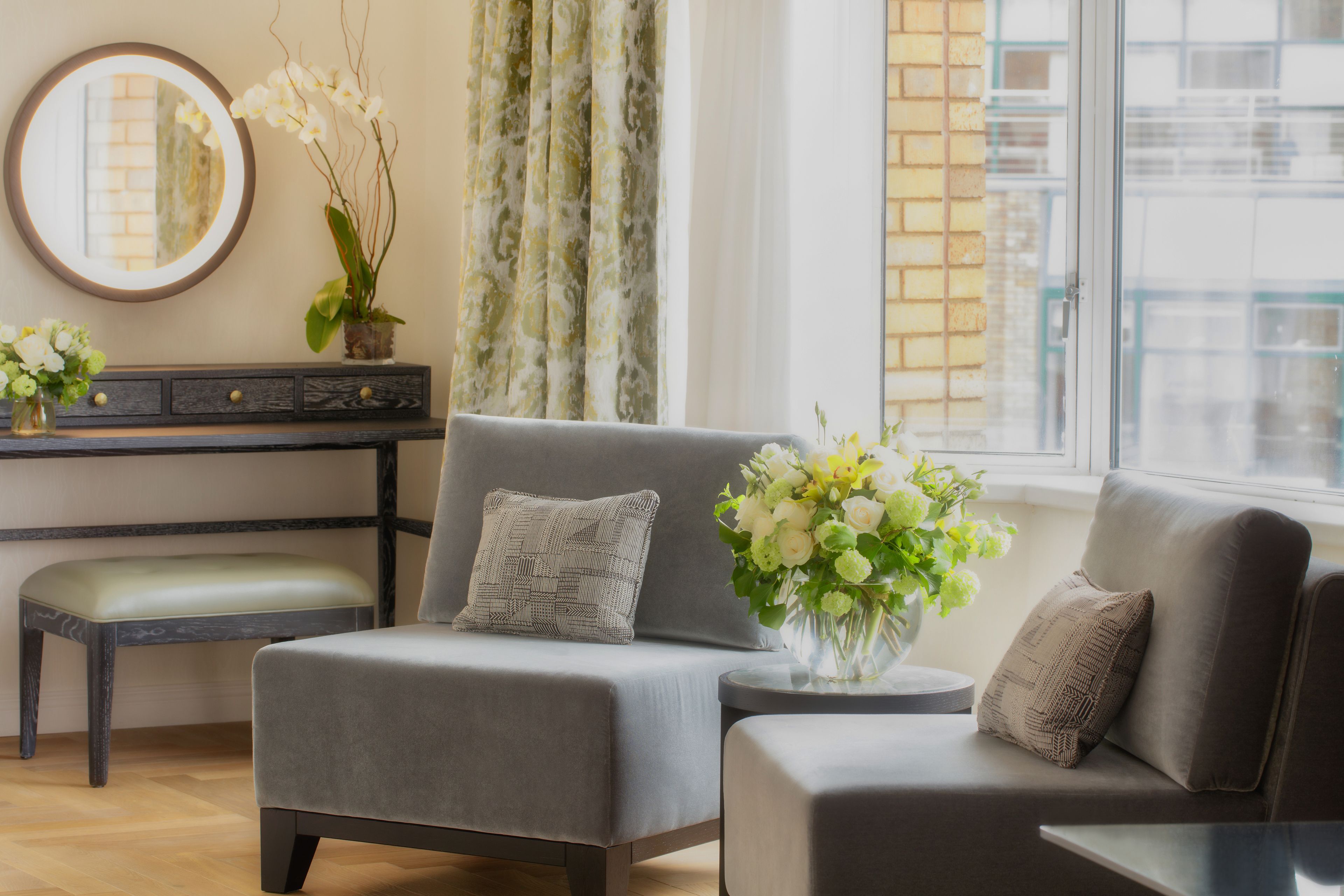 A modern living room with a grey sofa, a small table with a vase of flowers, a round mirror, and a window with curtains.