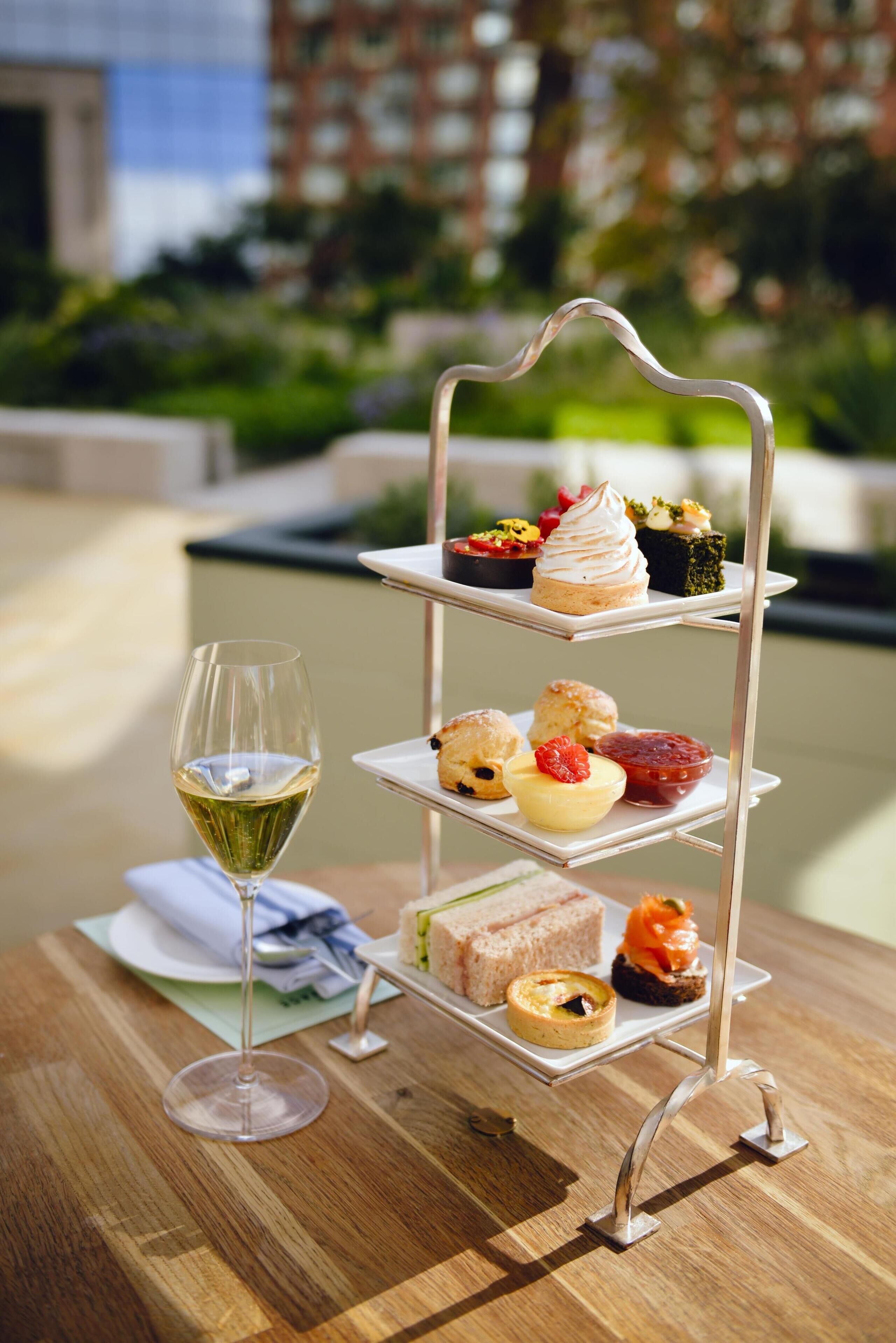 A tiered dessert stand with various pastries and a glass of white wine on a wooden table.