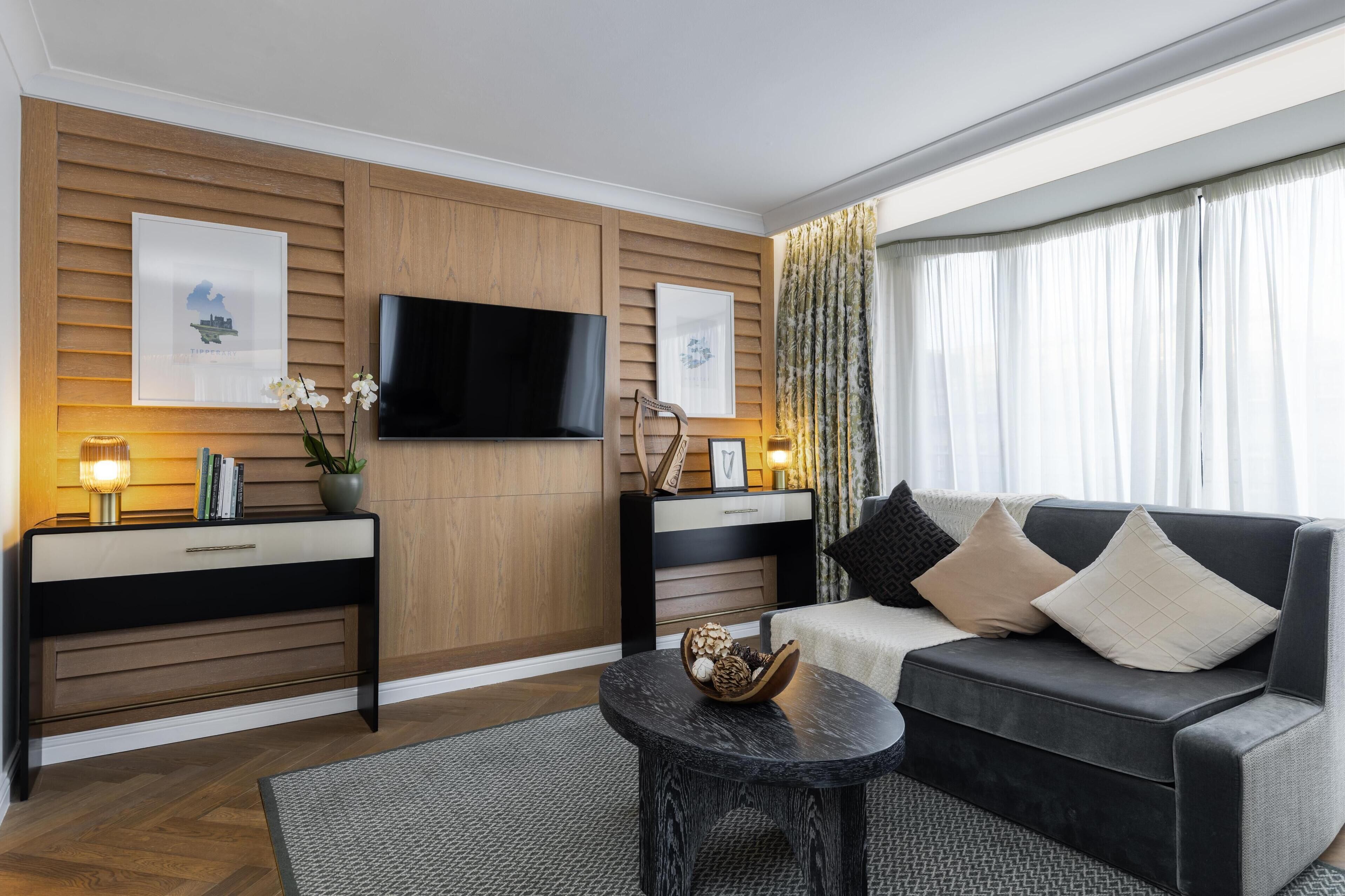 A modern living room with a flat-screen TV mounted on the wall, a sofa with cushions, a coffee table, and a side table with a lamp and decorative items.