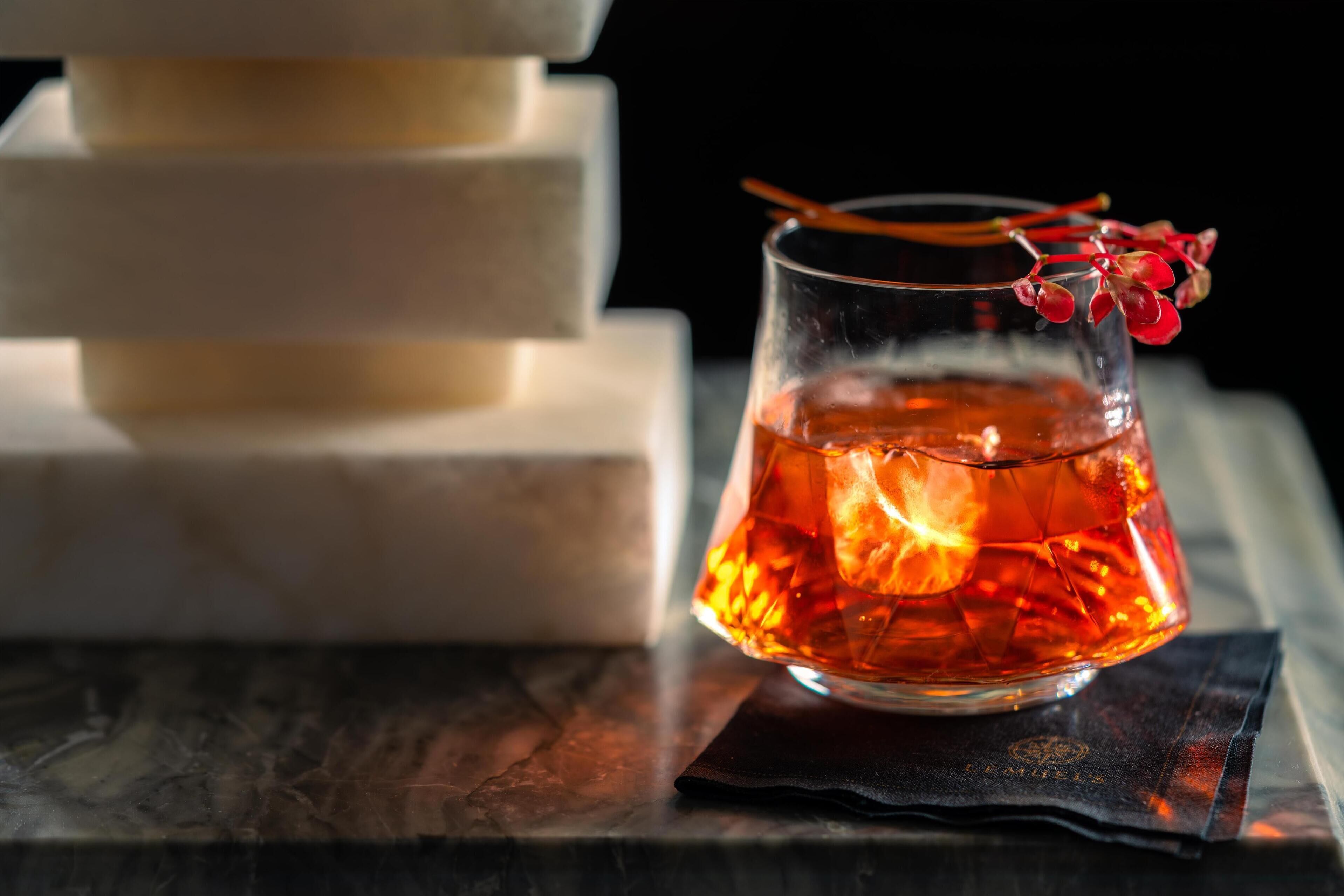 Un verre contenant un liquide ambré avec des glaçons, une tige décorative et une baie rouge en guise de décoration, le tout posé sur une serviette sombre.