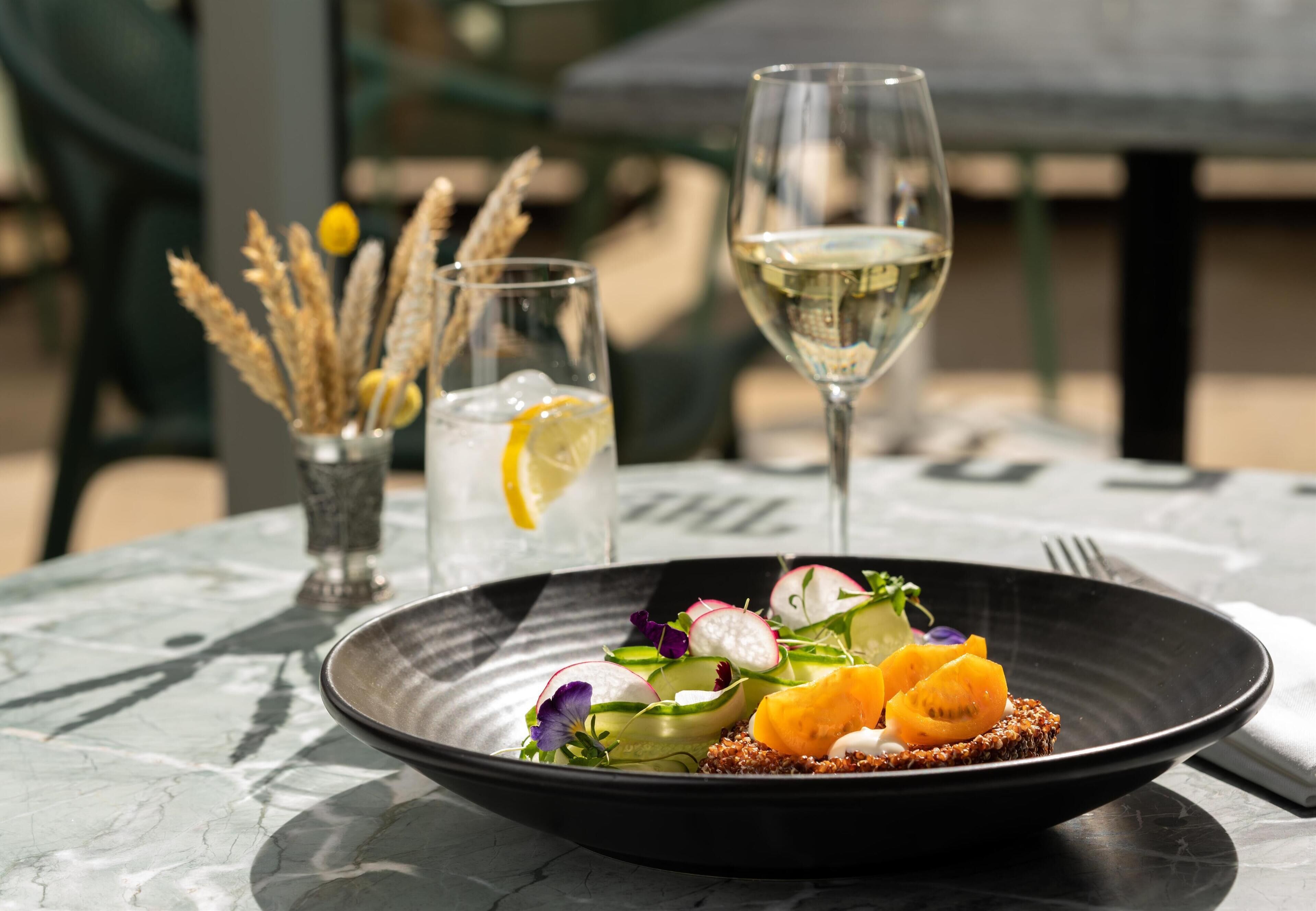 Una mesa de comedor con un plato negro de comida, una copa de vino blanco y un vaso de bebida helada con una rodaja de limón.